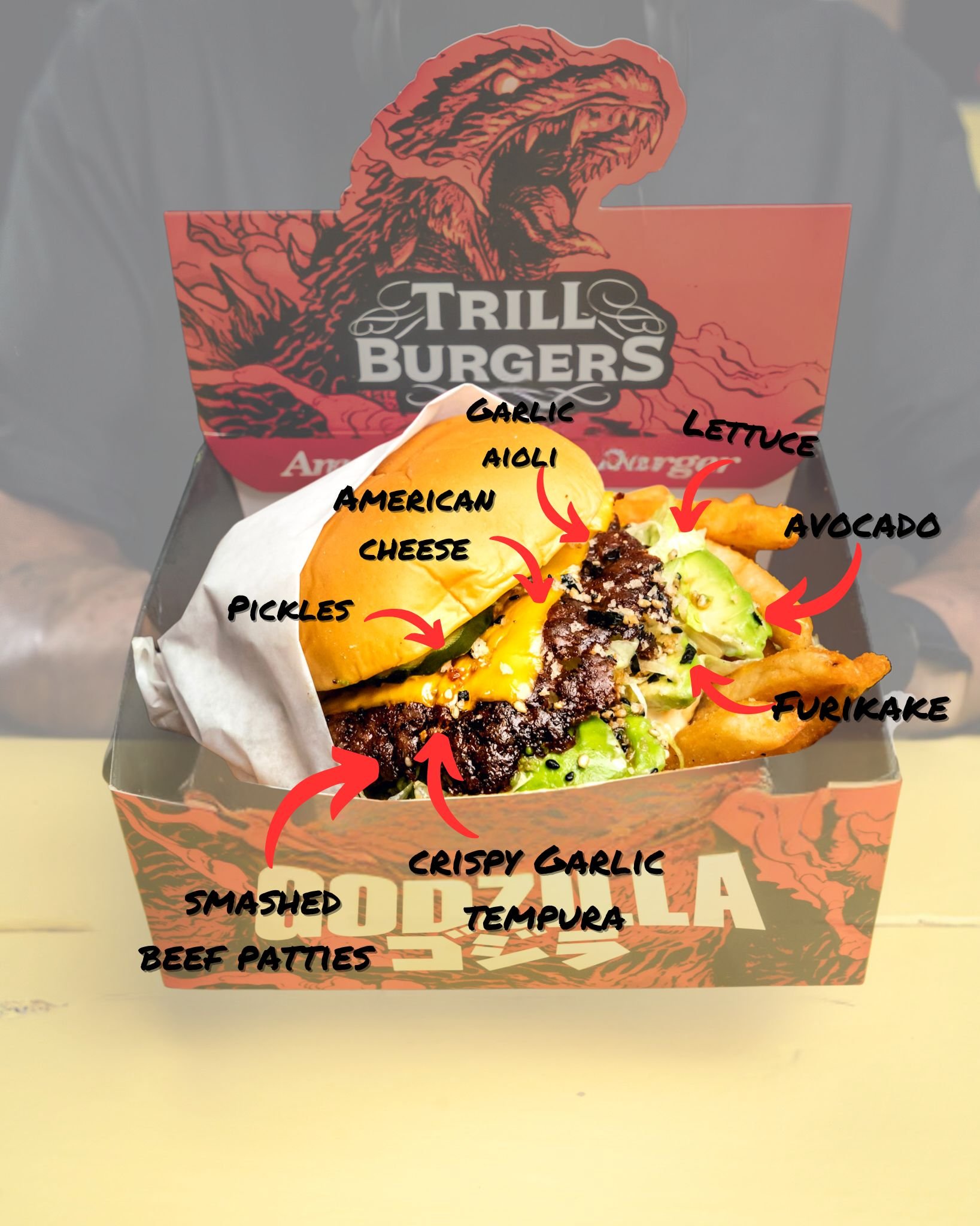 A burger with labeled ingredients including American cheese, pickles, smashed beef patties, crispy garlic, tempura, aioli, garlic, cheese, lettuce, avocado, and furikake in a branded box with a dinosaur logo.