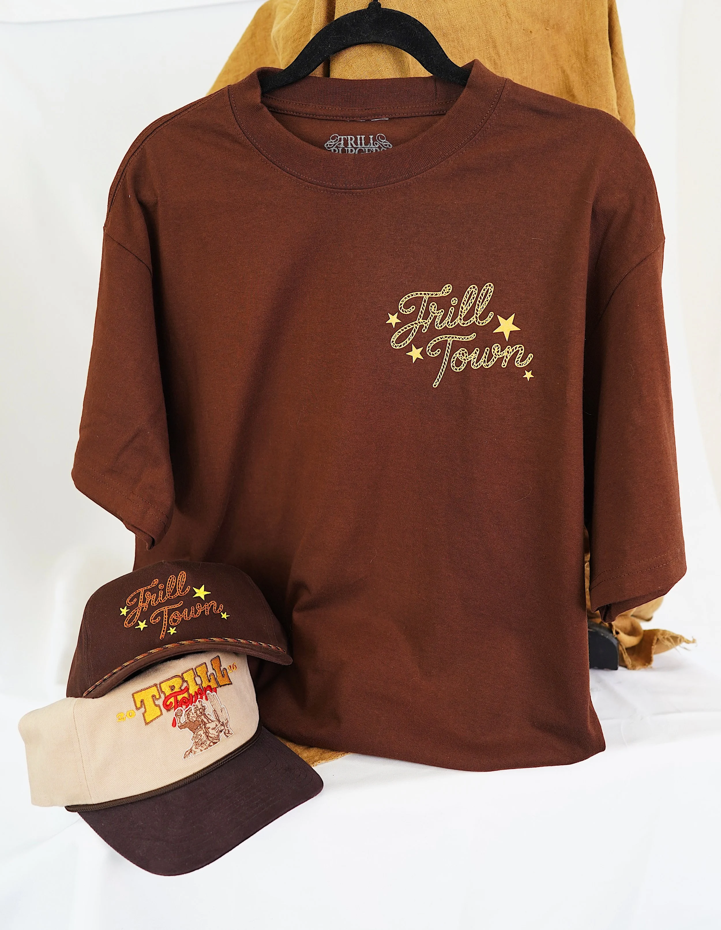 A brown T-shirt hanging on a black hanger with the phrase 'Trill Town' and stars in gold on the front. Two caps, one brown and one beige, both with embroidered 'Trill Town' logos, are placed in front of the T-shirt.