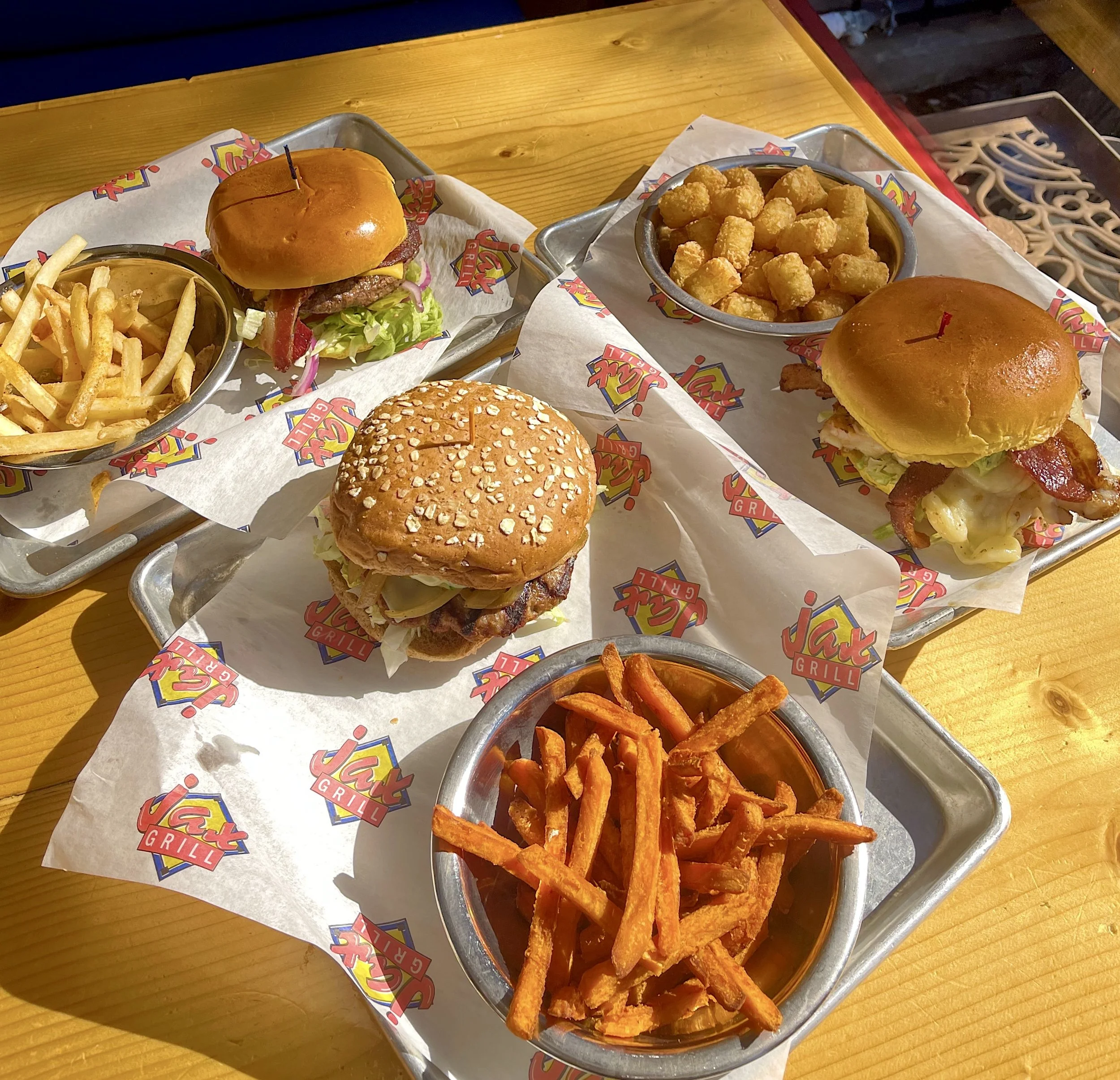 Three trays with burgers, fries, tater tots, and sweet potato fries on a wooden table at Jack's Grill.