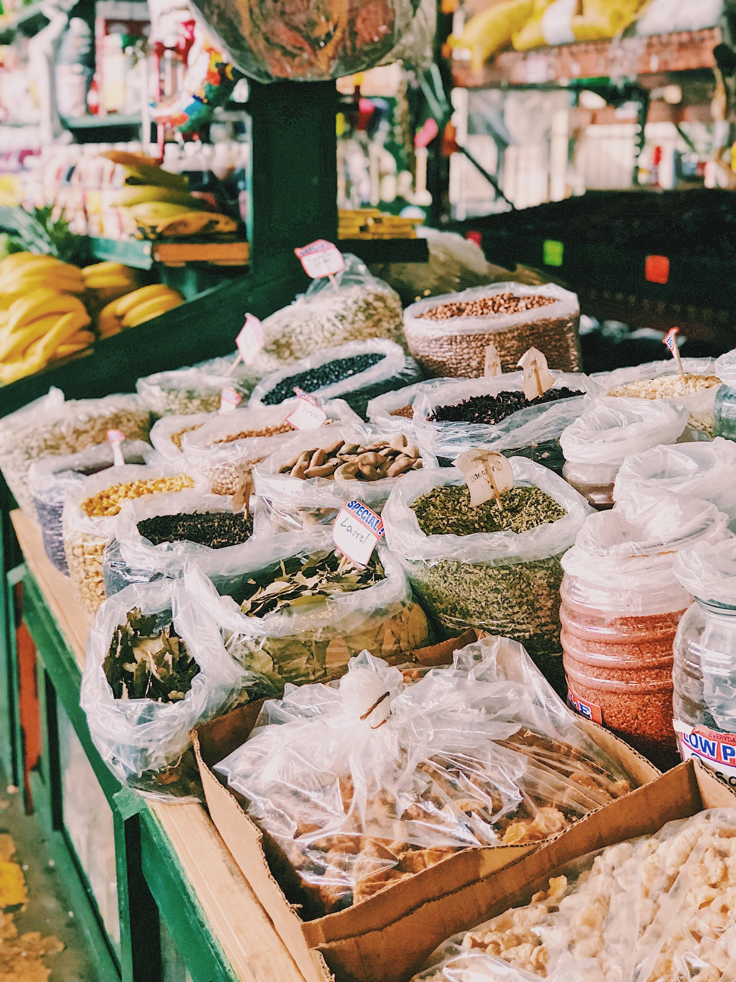 Various dried herbs and spices in clear plastic bags on a market stand, with bananas and other produce in the background.
