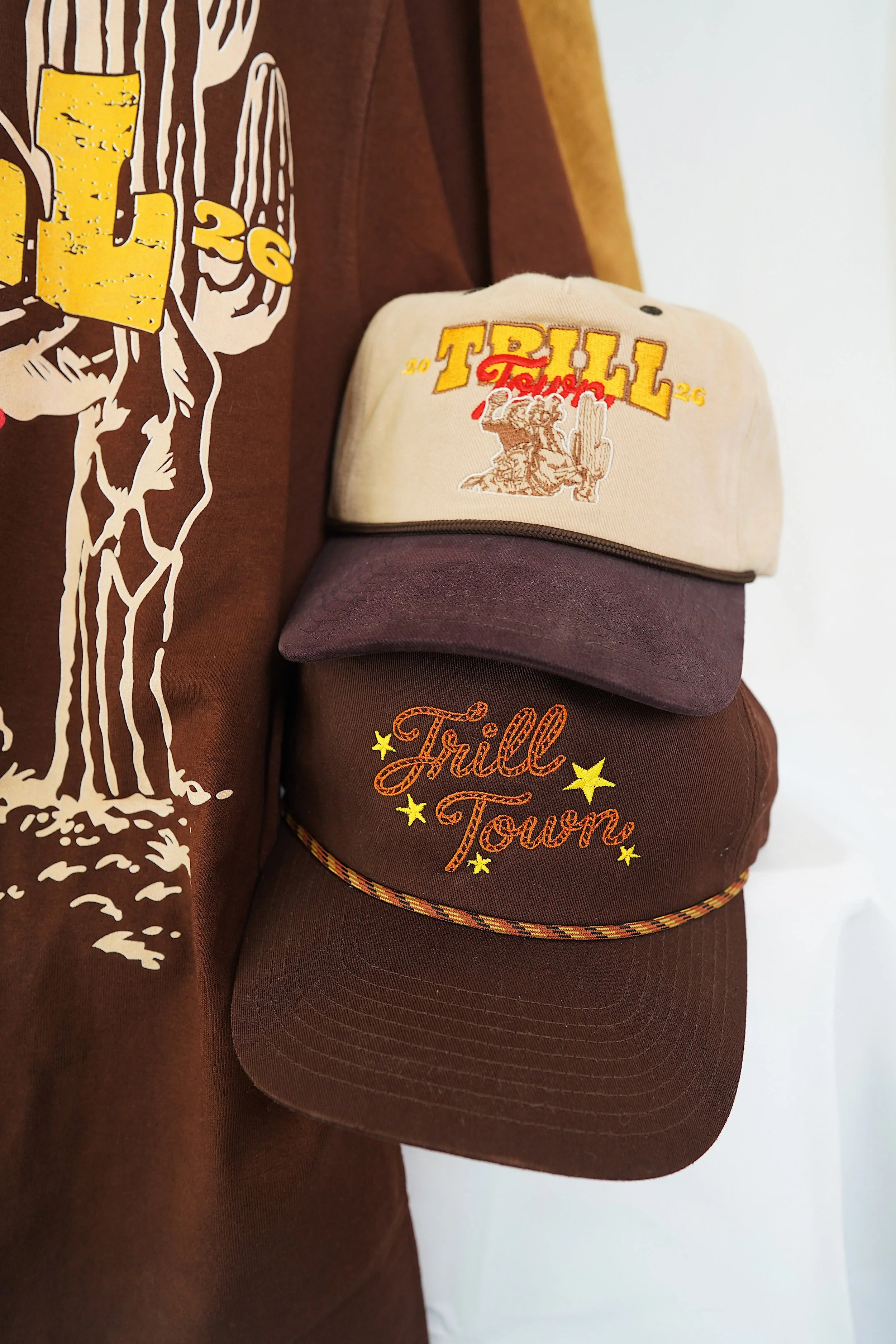 Stack of three hats hanging on a brown fabric. The top hat is cream-colored with yellow and red embroidery that says 'TRILL 2026.' The middle hat is plain dark purple. The bottom hat is brown with orange embroidery that says 'Trill Town' with yellow 