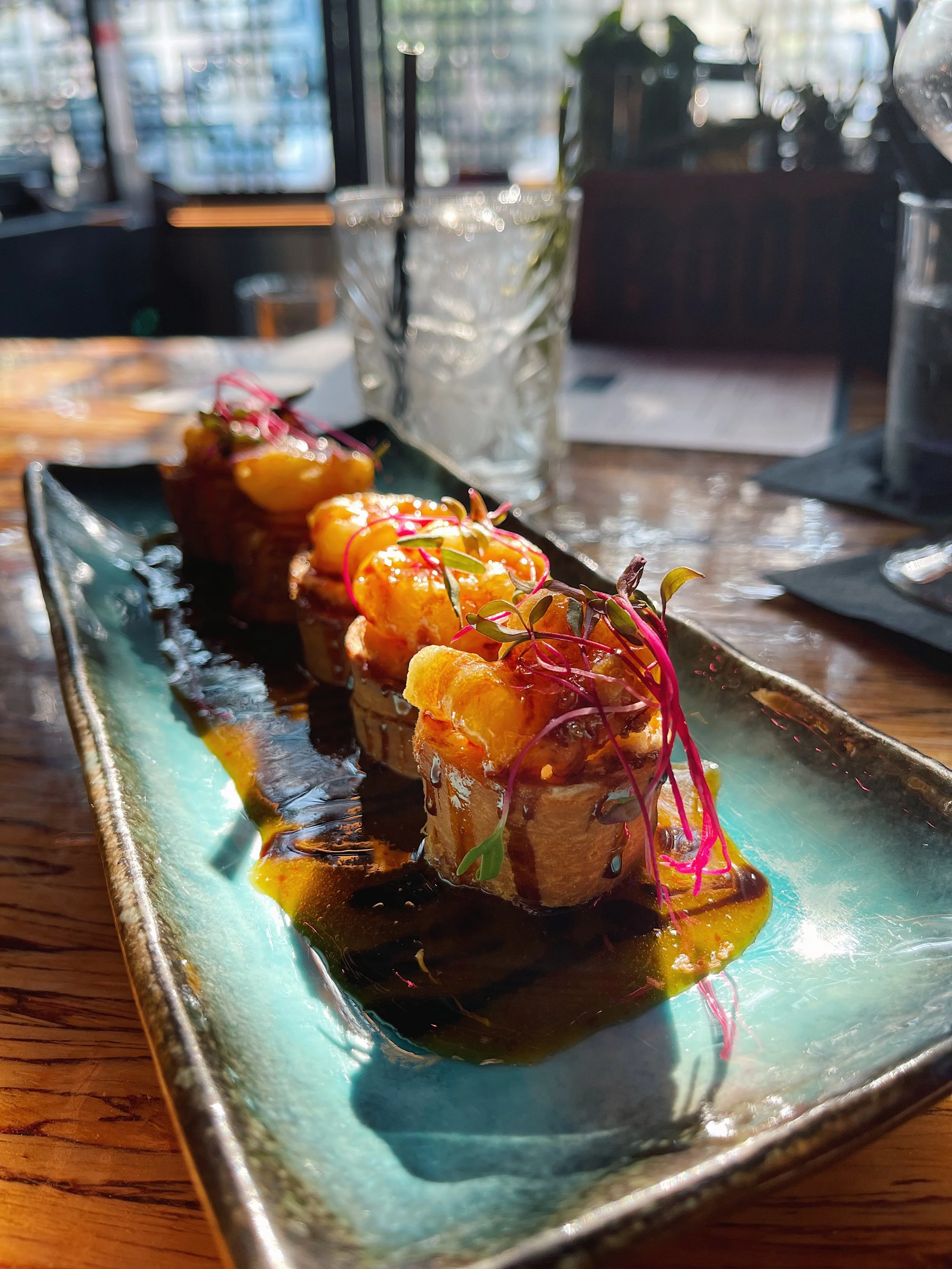 Close-up of an elegant, multi-piece sushi dish with bright toppings, garnished with microgreens and drizzled with sauce, served on a rectangular ceramic plate, set on a wooden table in a restaurant.