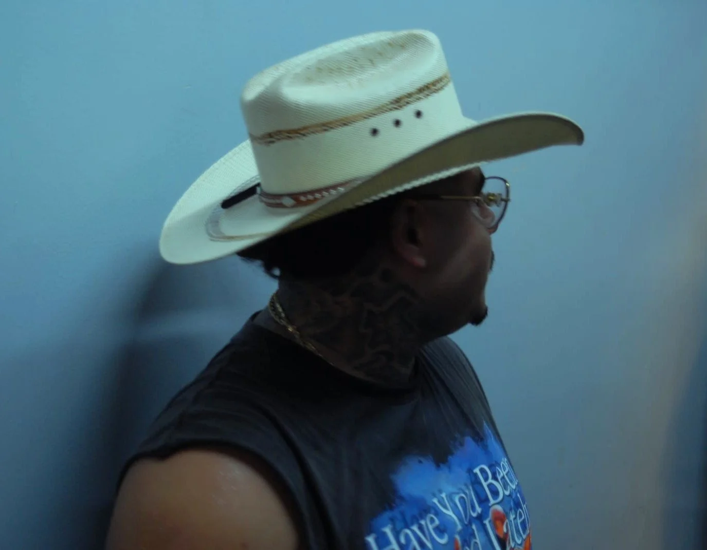 A person wearing glasses, a dark tie-dye shirt, and a large white cowboy hat with a decorative band, standing against a plain light blue wall.