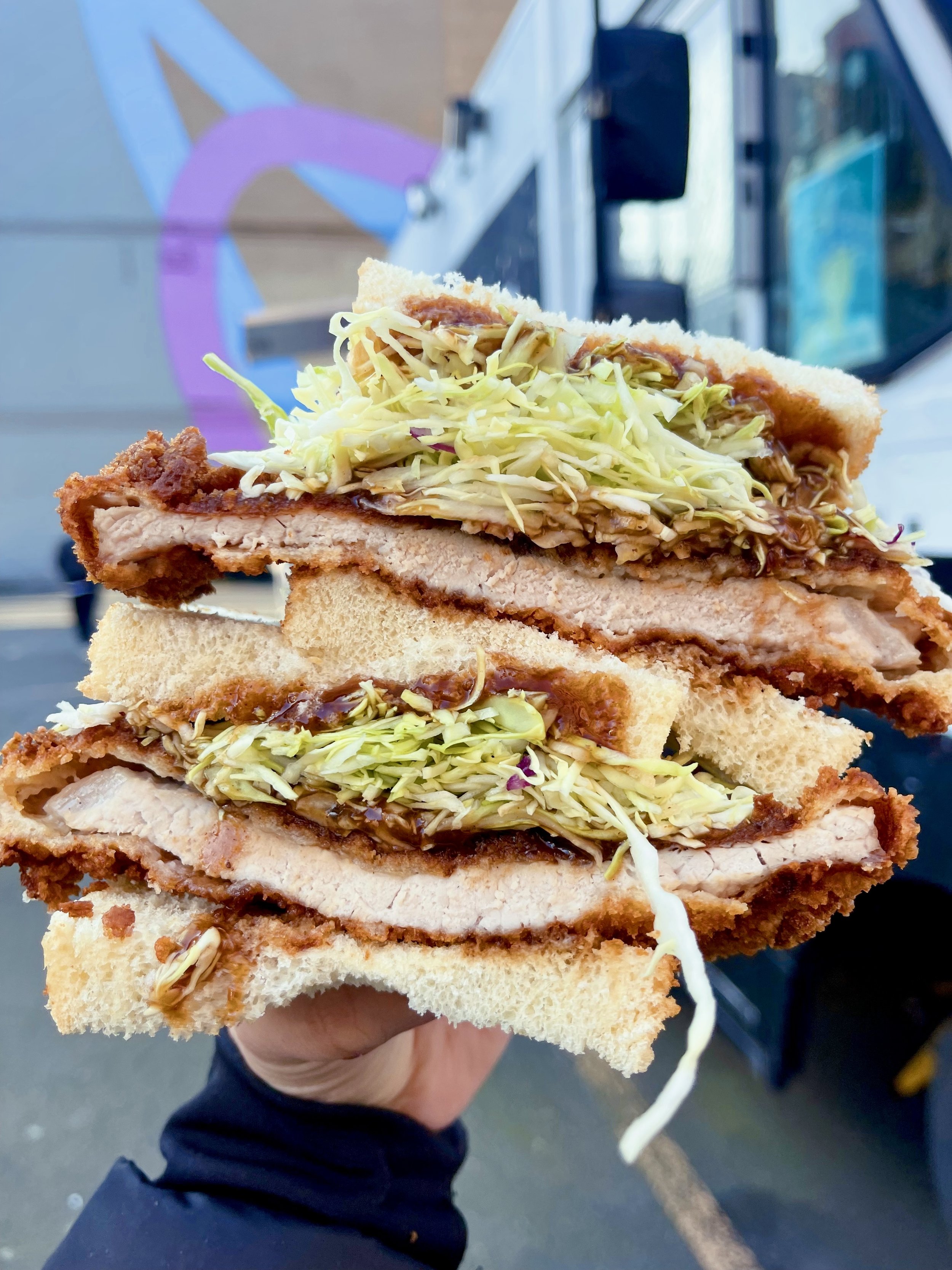 Close-up of a fried chicken sandwich with shredded lettuce and sauce in a bun, held outside near a food truck.