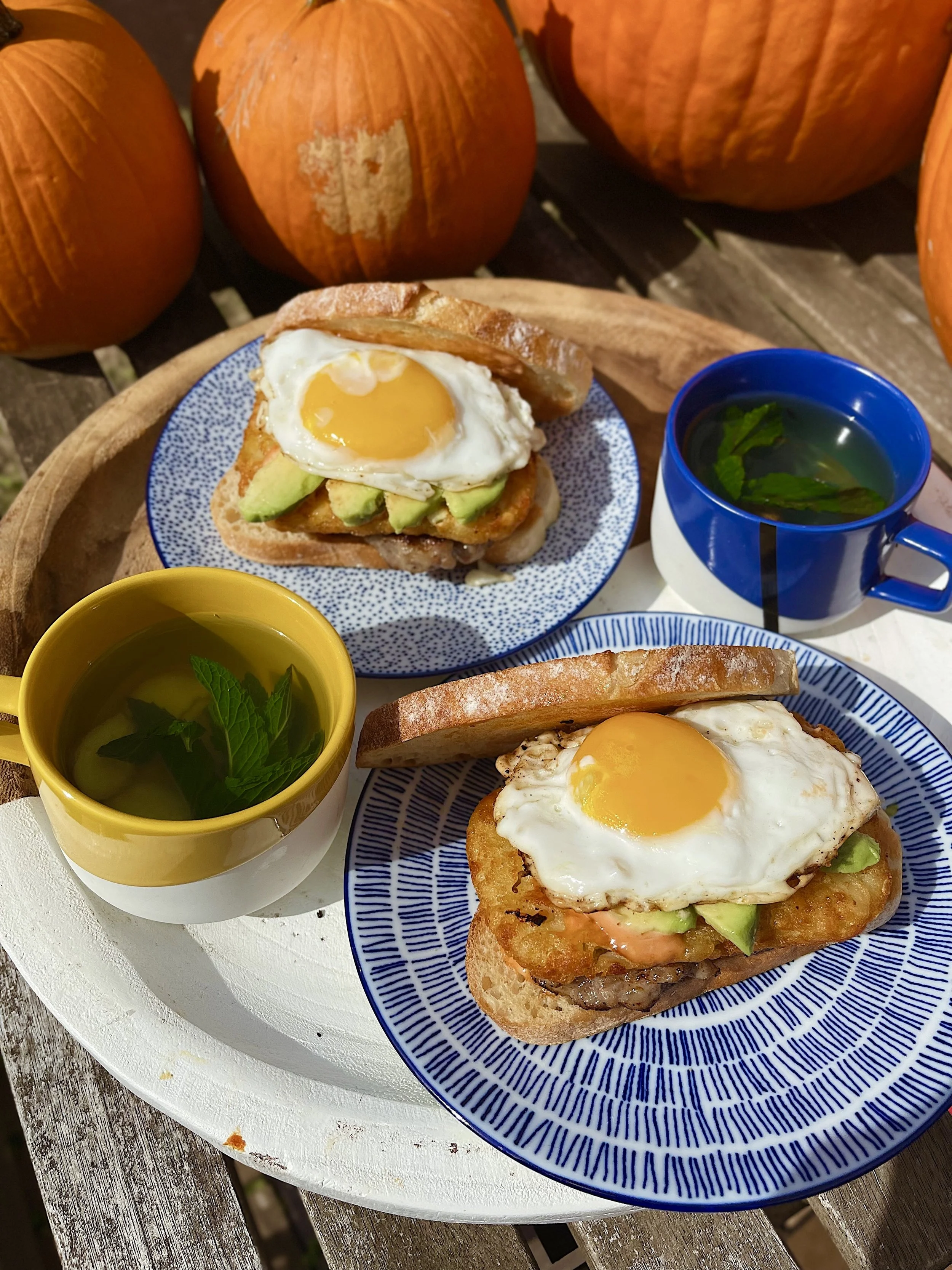 Two breakfast sandwiches with fried eggs, avocado slices, and bacon in toasted buns, served on patterned plates. There are two cups of tea with mint leaves and a cup with herbs, all on a white tray. In the background, there are three pumpkins.