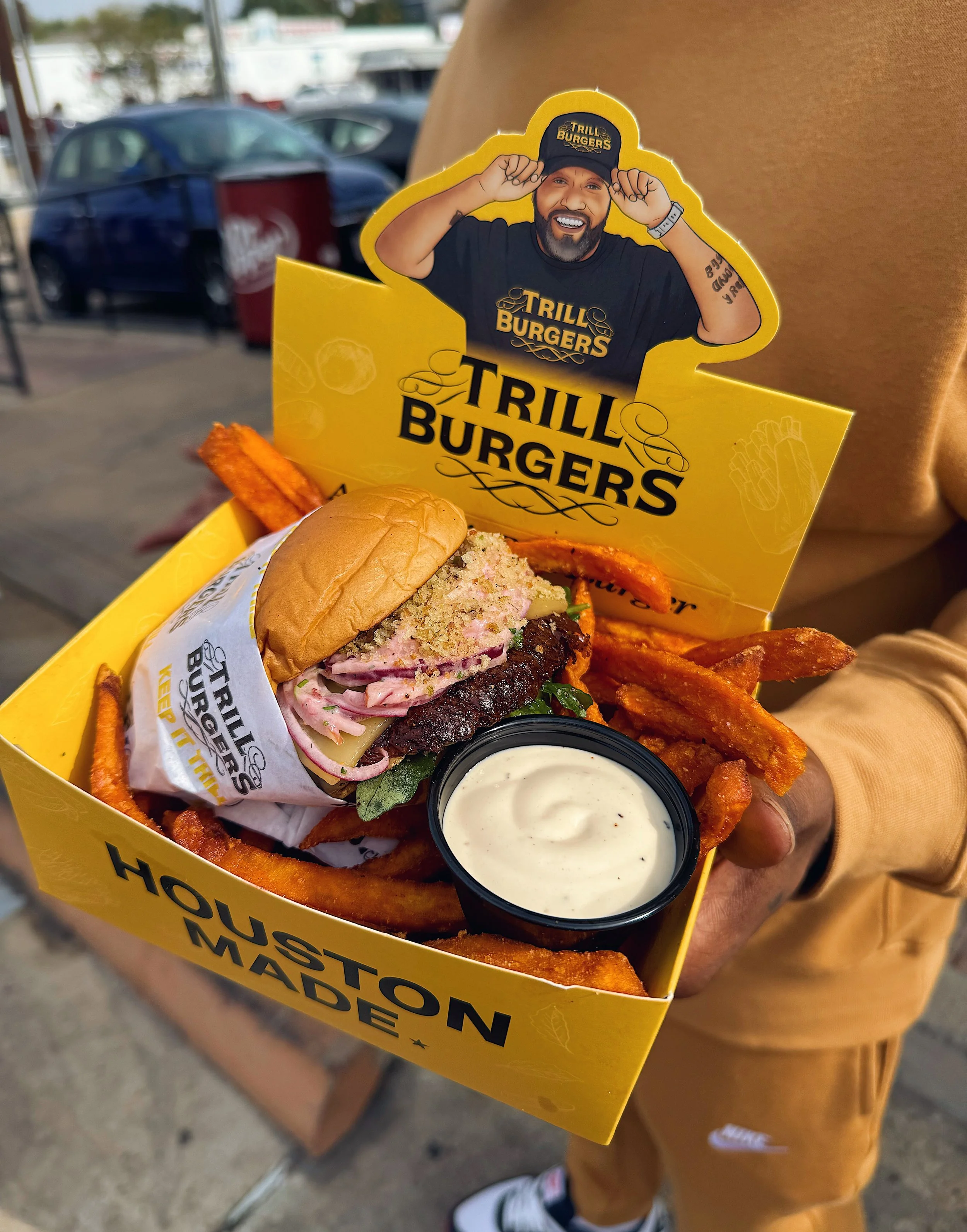 A person holding a yellow box of food from Houston Made with fries, a burger with coleslaw, and a side of ranch dressing. The box has a cardboard cutout of a man wearing a Trill Burgers hat and shirt, smiling and pulling on his hat.
