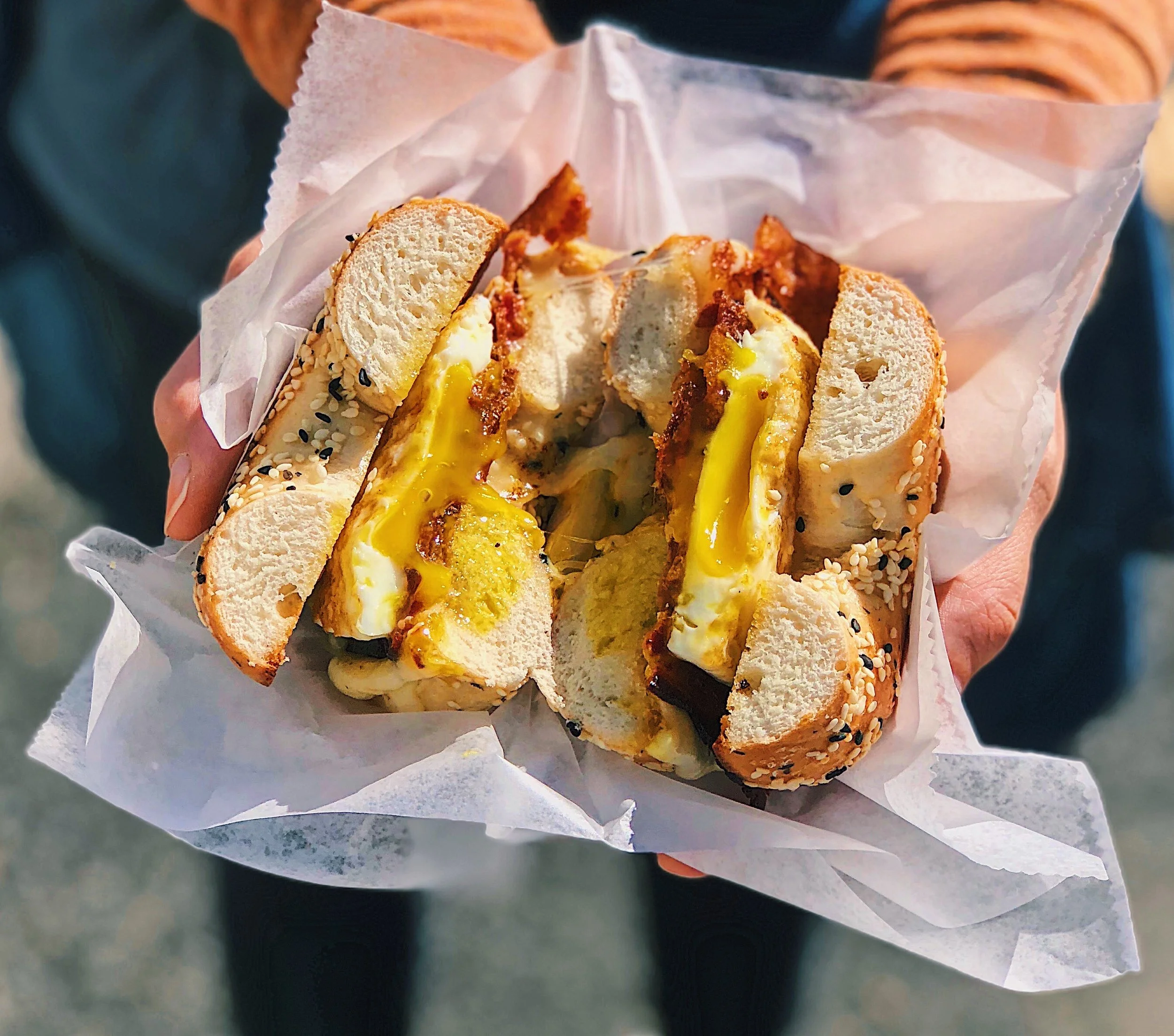 A person holding a sandwich with egg, bacon, and bread, wrapped in white paper.