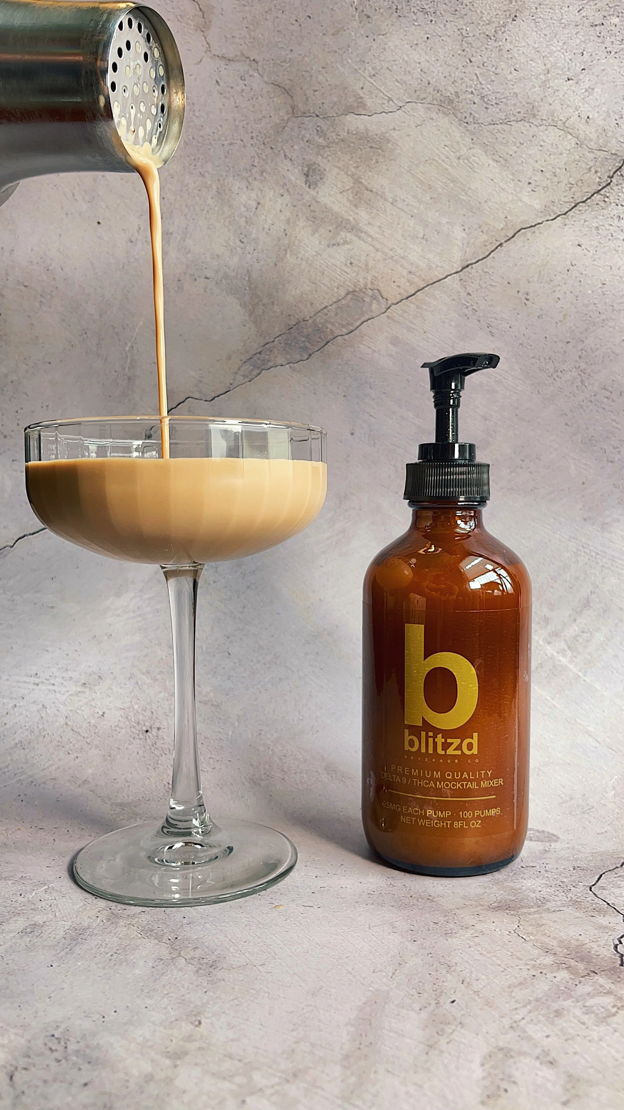A glass with a stem being filled with a beige liquid from a canister, placed on a textured gray surface. A brown bottle labeled 'blitz' with a pump dispenser is next to the glass.