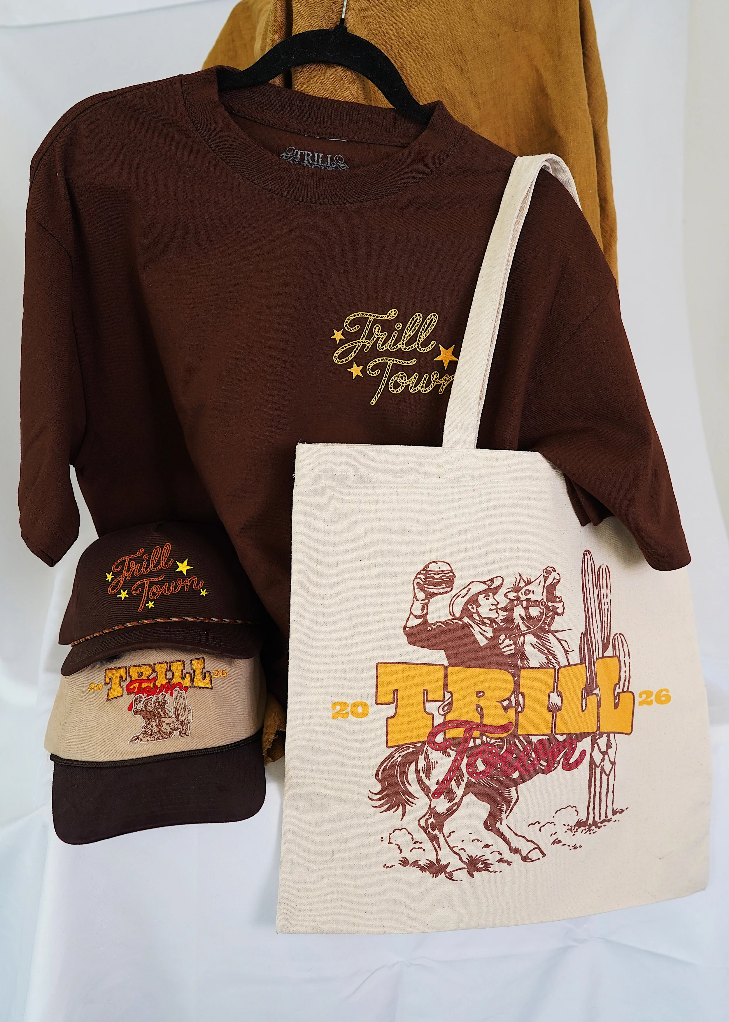 Collection of 'Fill Town 26' merchandise including a brown t-shirt, a cap, and a tote bag with western-themed graphics and text.
