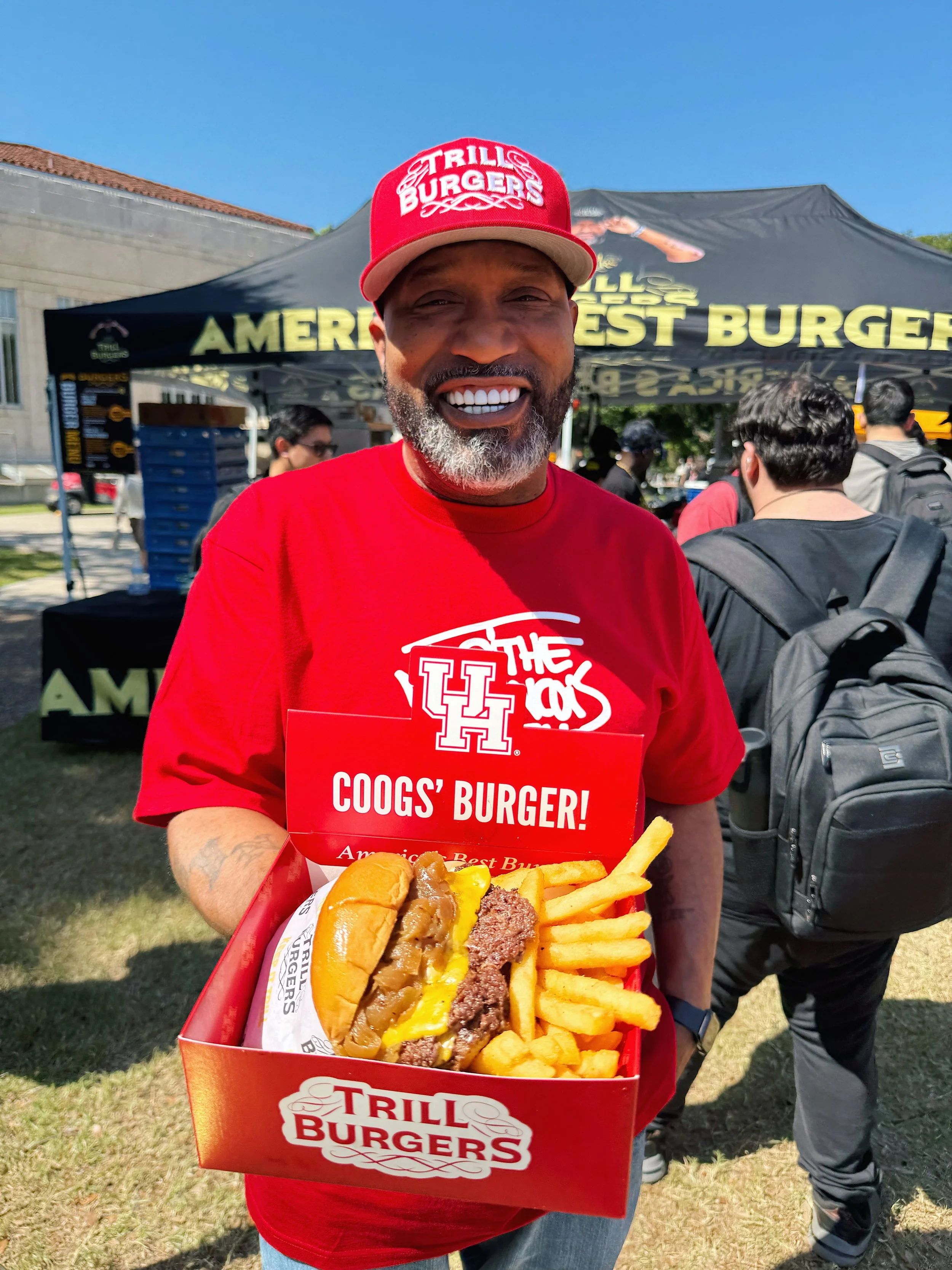 Man smiling and holding a tray with a burger and fries at a food festival with a Trill Burgers tent in the background.