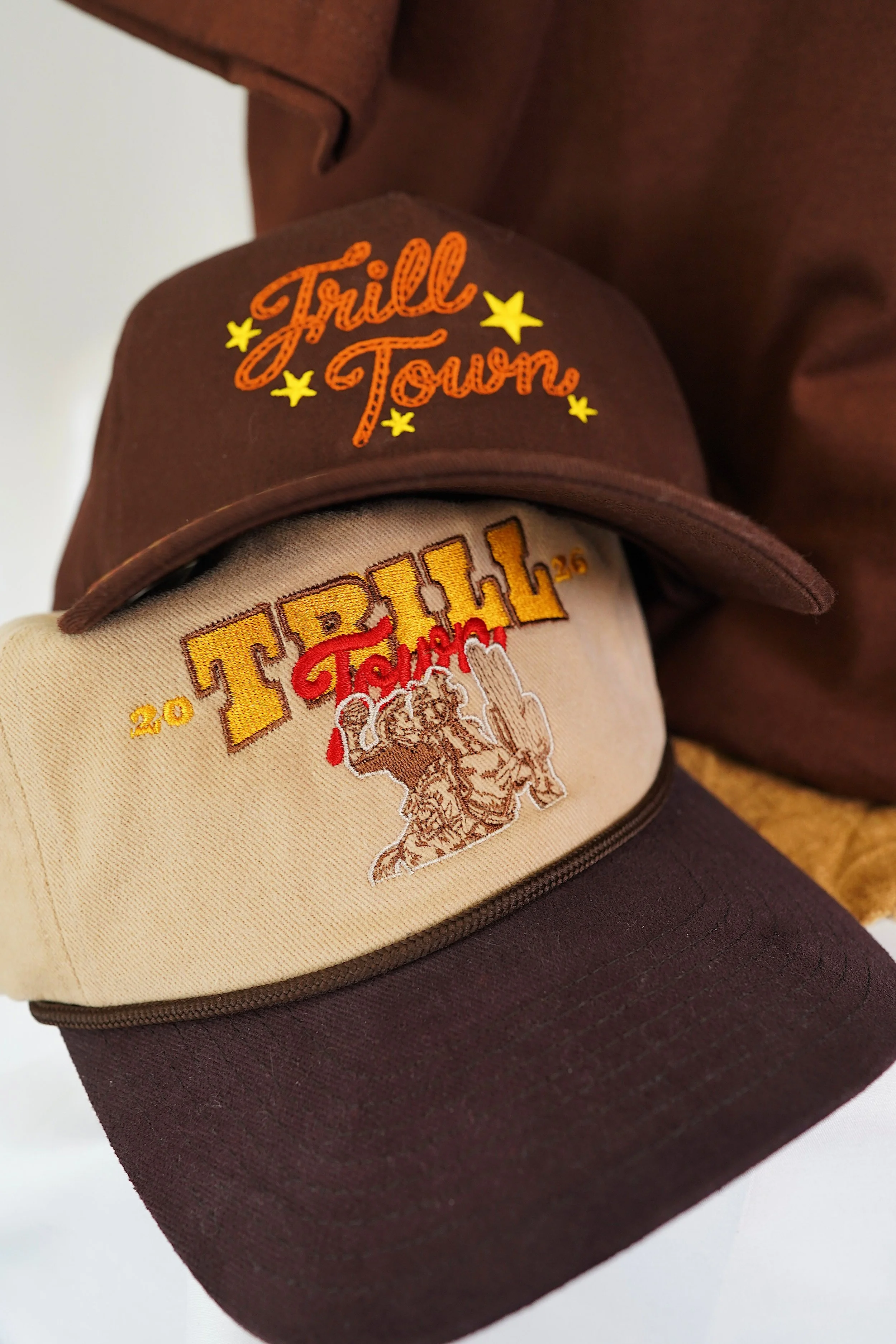 A close-up of two caps, one beige and one dark brown, featuring embroidered text and graphics. The beige cap has "TRILL" in yellow and a drawing of a lion, while the dark brown cap has "Trill Town" in orange and yellow stars.