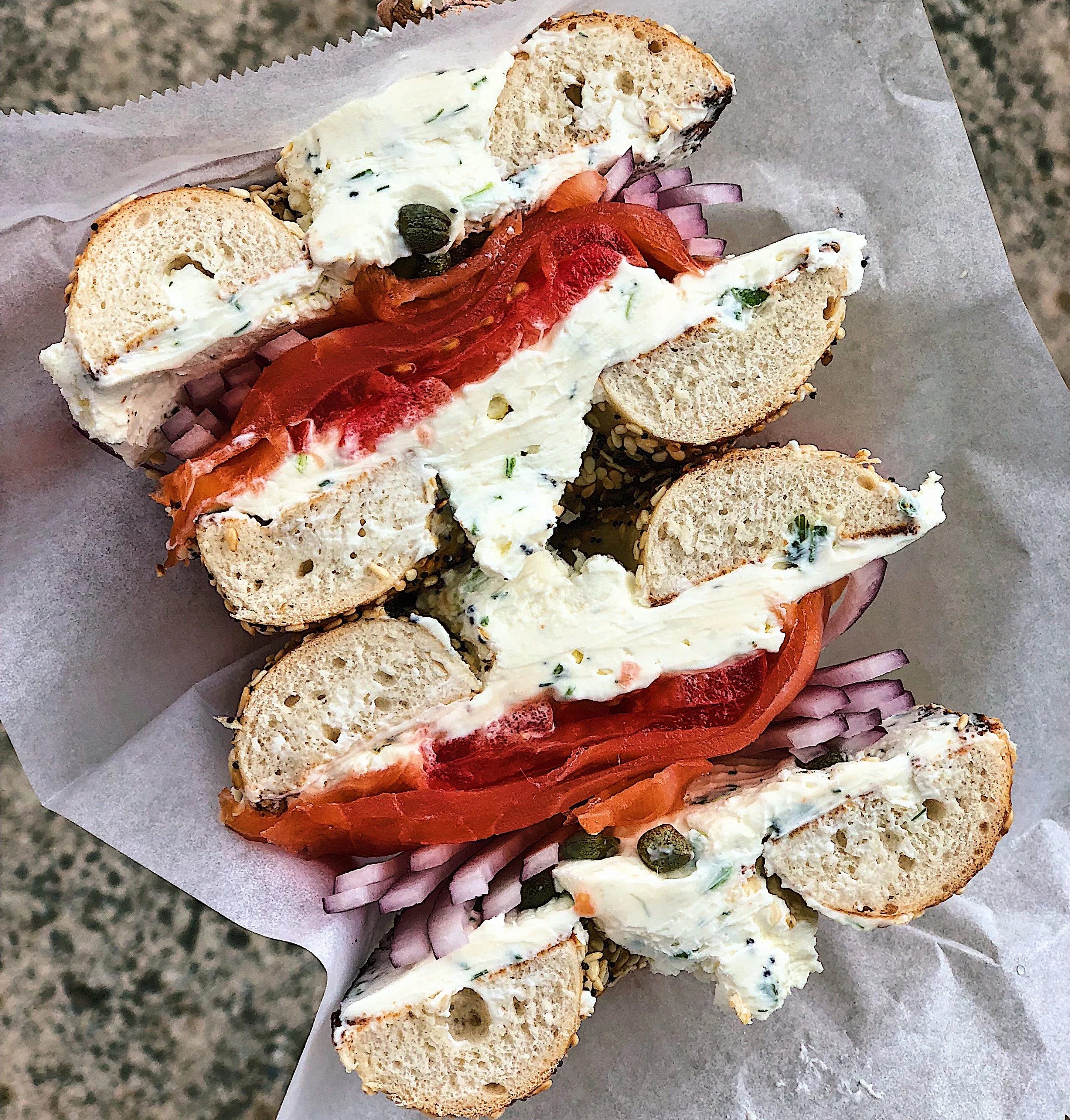 A bagel sandwich with cream cheese, sliced tomato, red onion, capers, and smoked salmon, cut in half and served on parchment paper.
