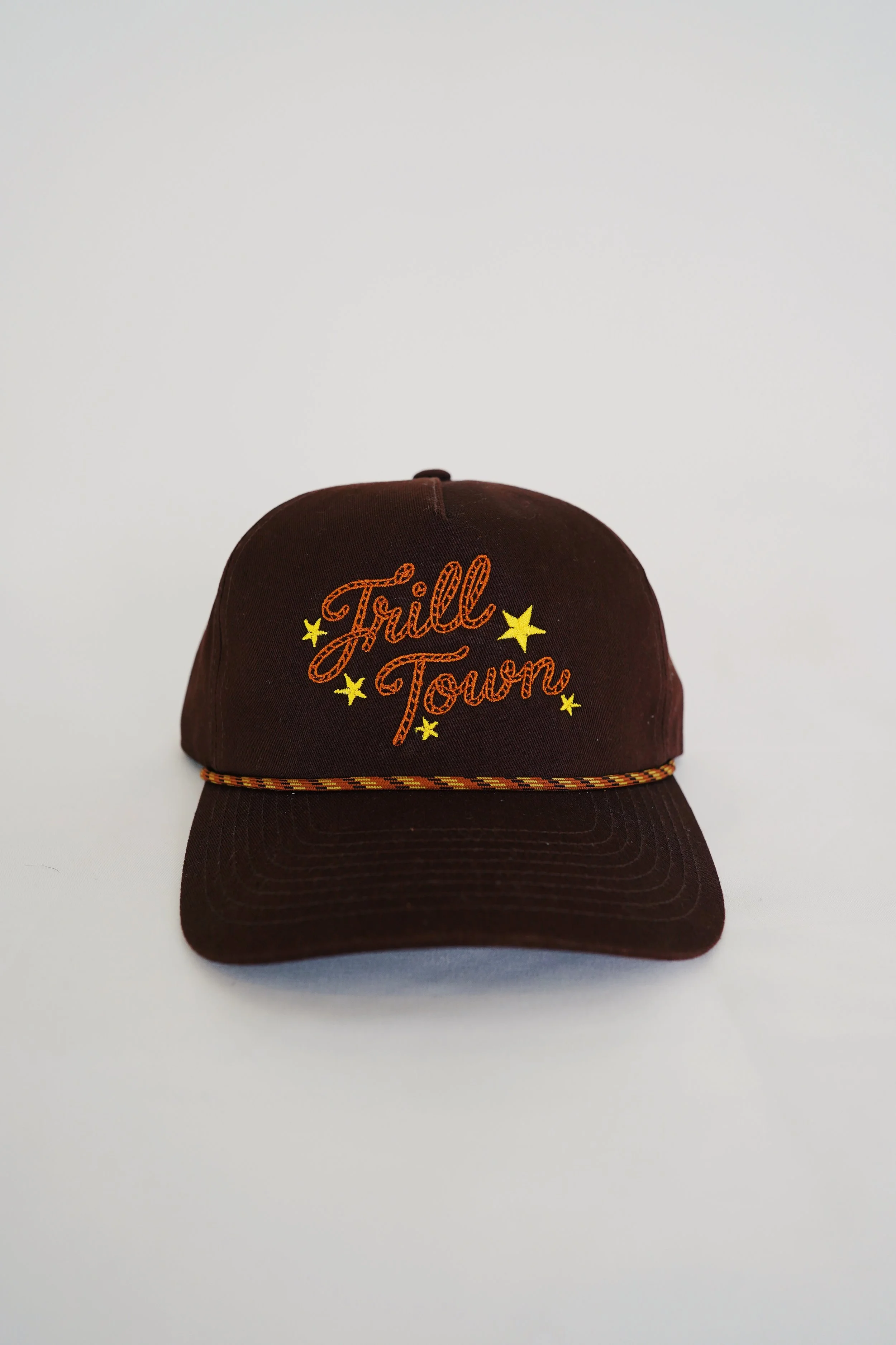 A dark brown baseball cap with embroidered orange text saying 'Trill Town' and yellow stars, sitting on a white background.