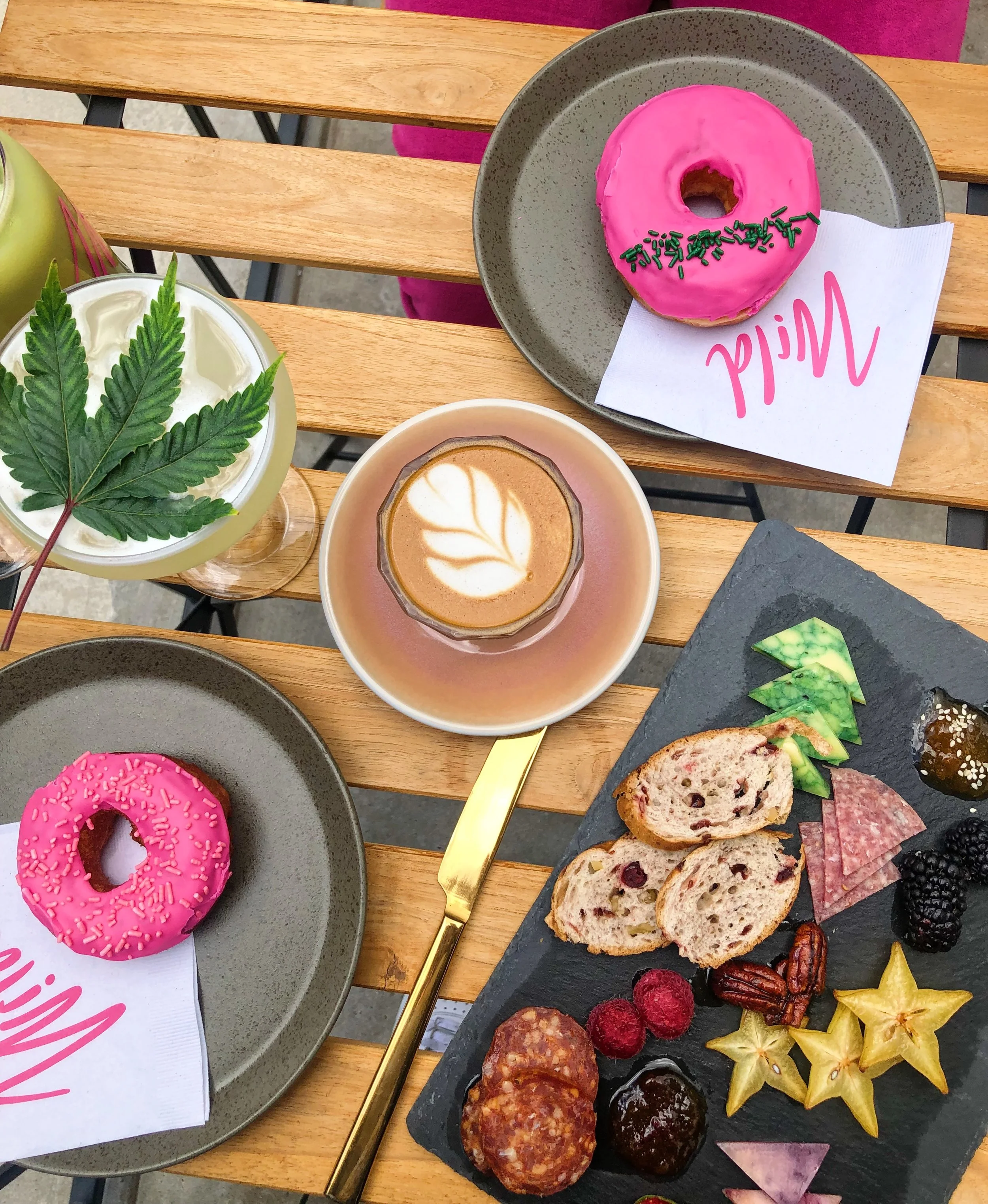 A wooden table set with two pink-glazed donuts with sprinkles on gray plates, a cappuccino with leaf latte art, a glass with whipped cream and a cannabis leaf, and a black slate platter with assorted bread, fruits, and snacks, all arranged for a brun