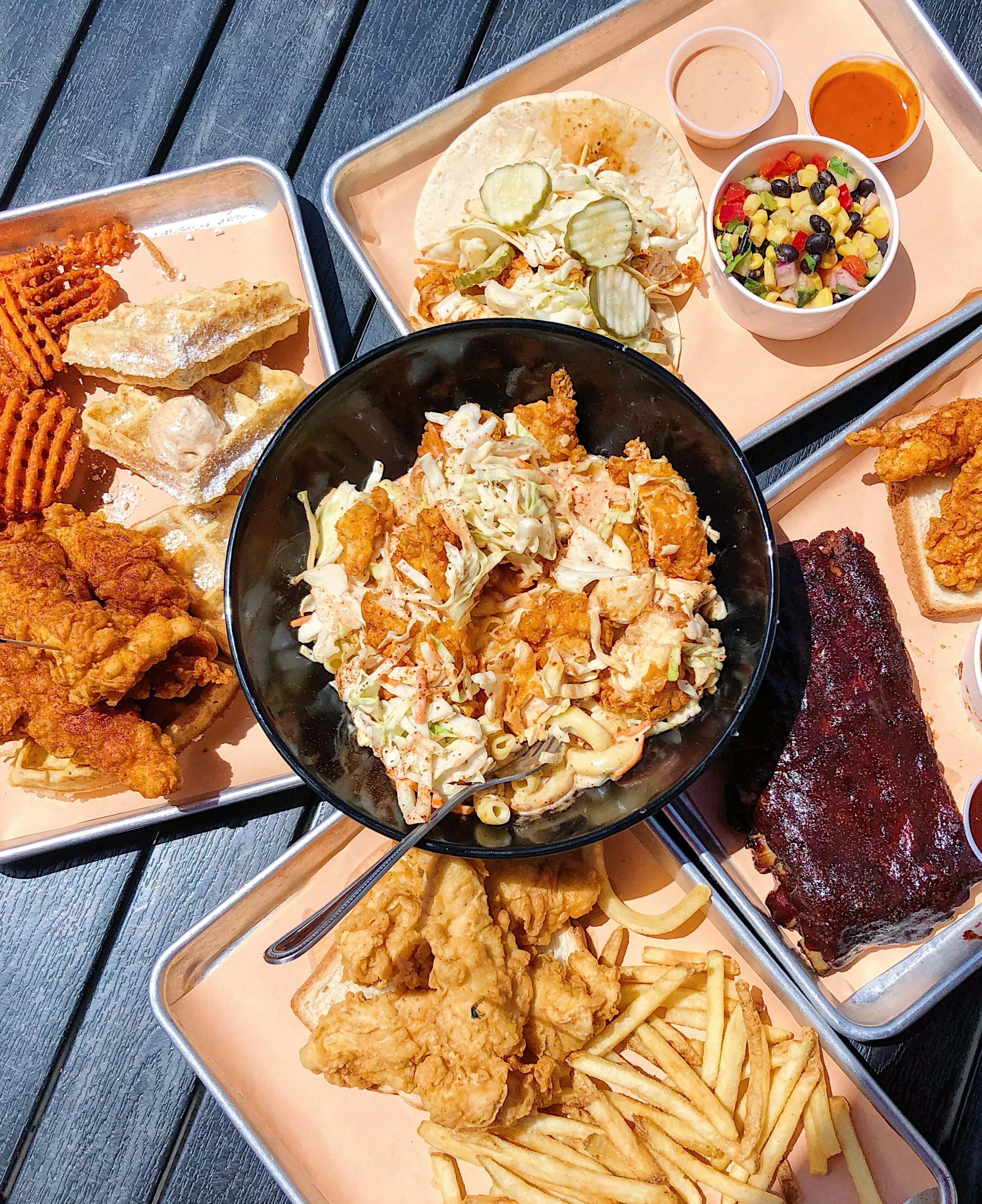 A variety of fried chicken, ribs, French fries, coleslaw, potato chips, pickles, salad, and dipping sauces on metal trays.