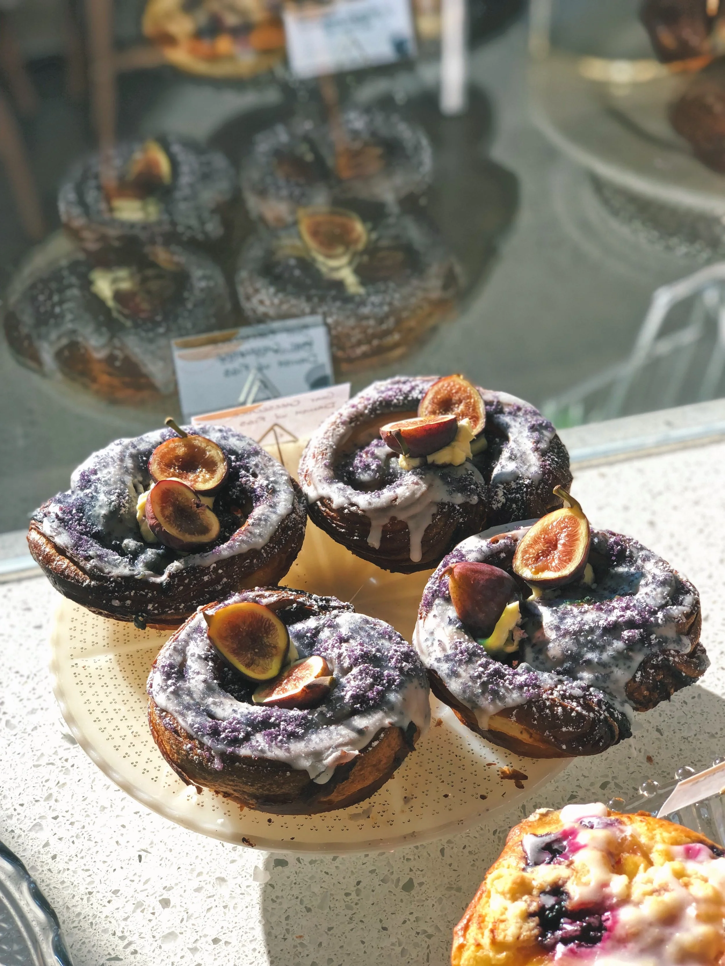 A plate of four chocolate cupcakes topped with purple and white icing, fresh halved figs, and purple sprinkles, displayed in a bakery window with other baked goods in the background.
