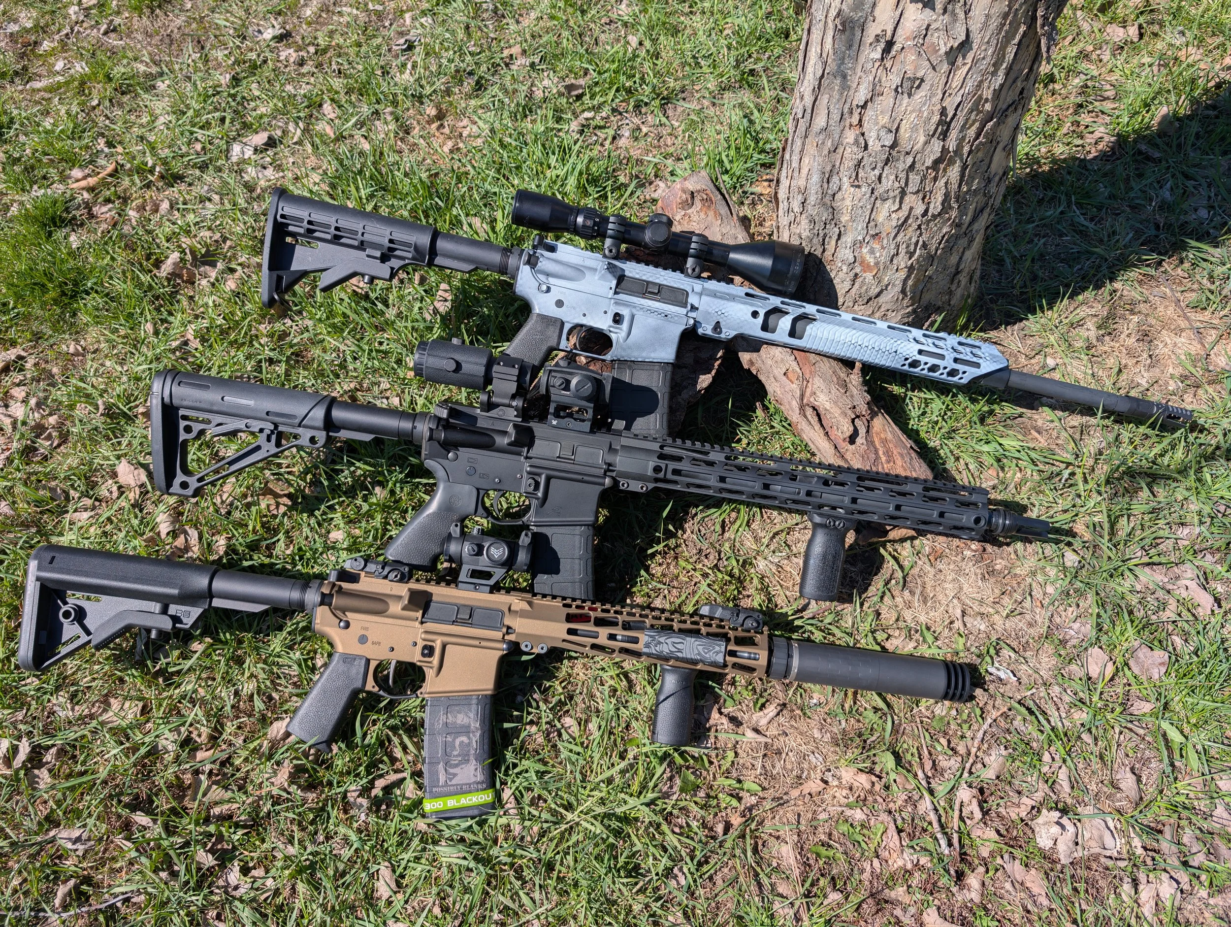 Three rifles with various attachments are resting on the grass near a tree trunk.