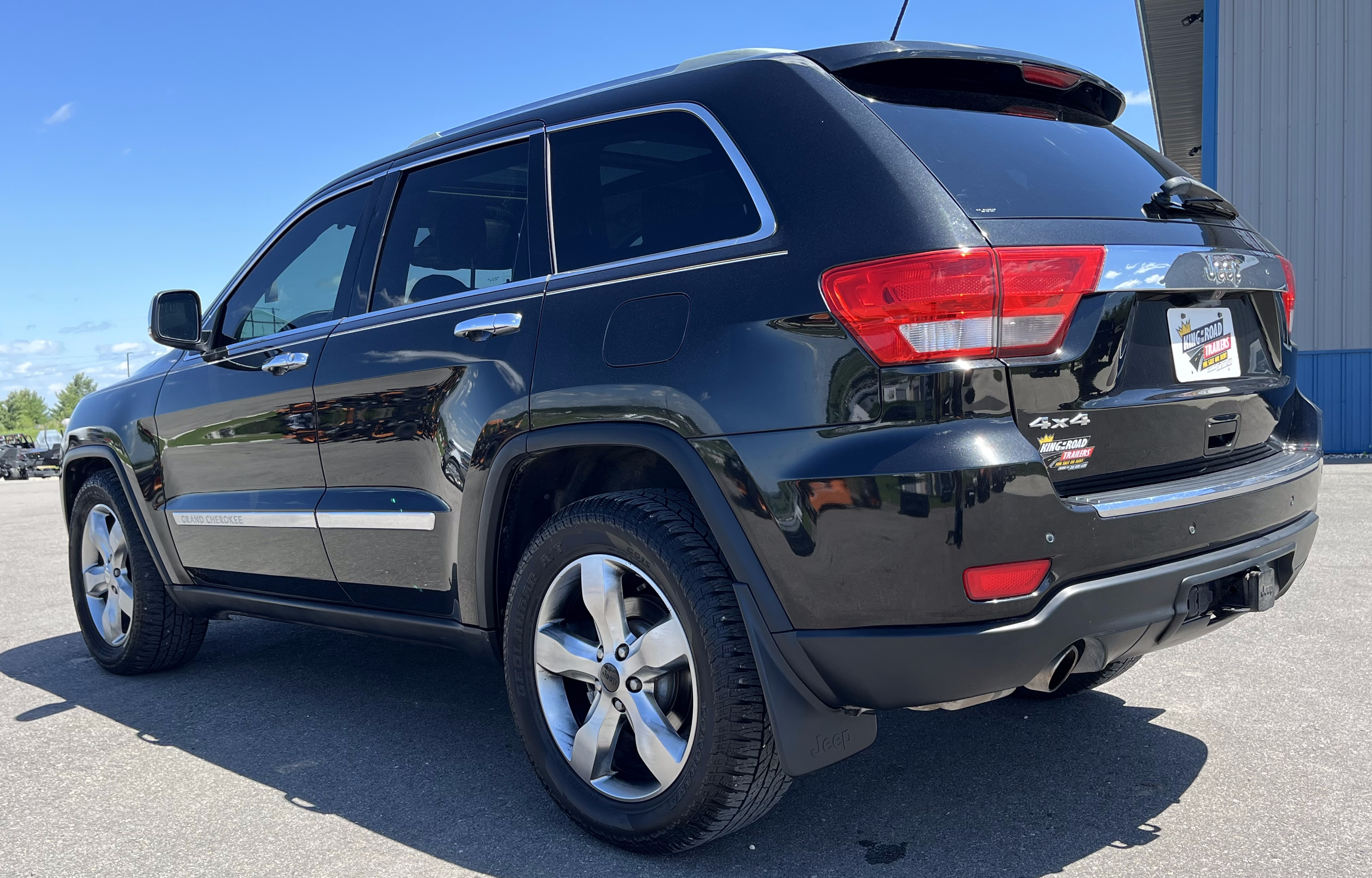 Black Jeep Grand Cherokee parked outdoors on a sunny day, showing the rear and side views, with a dealership plate and a trailer hitch.