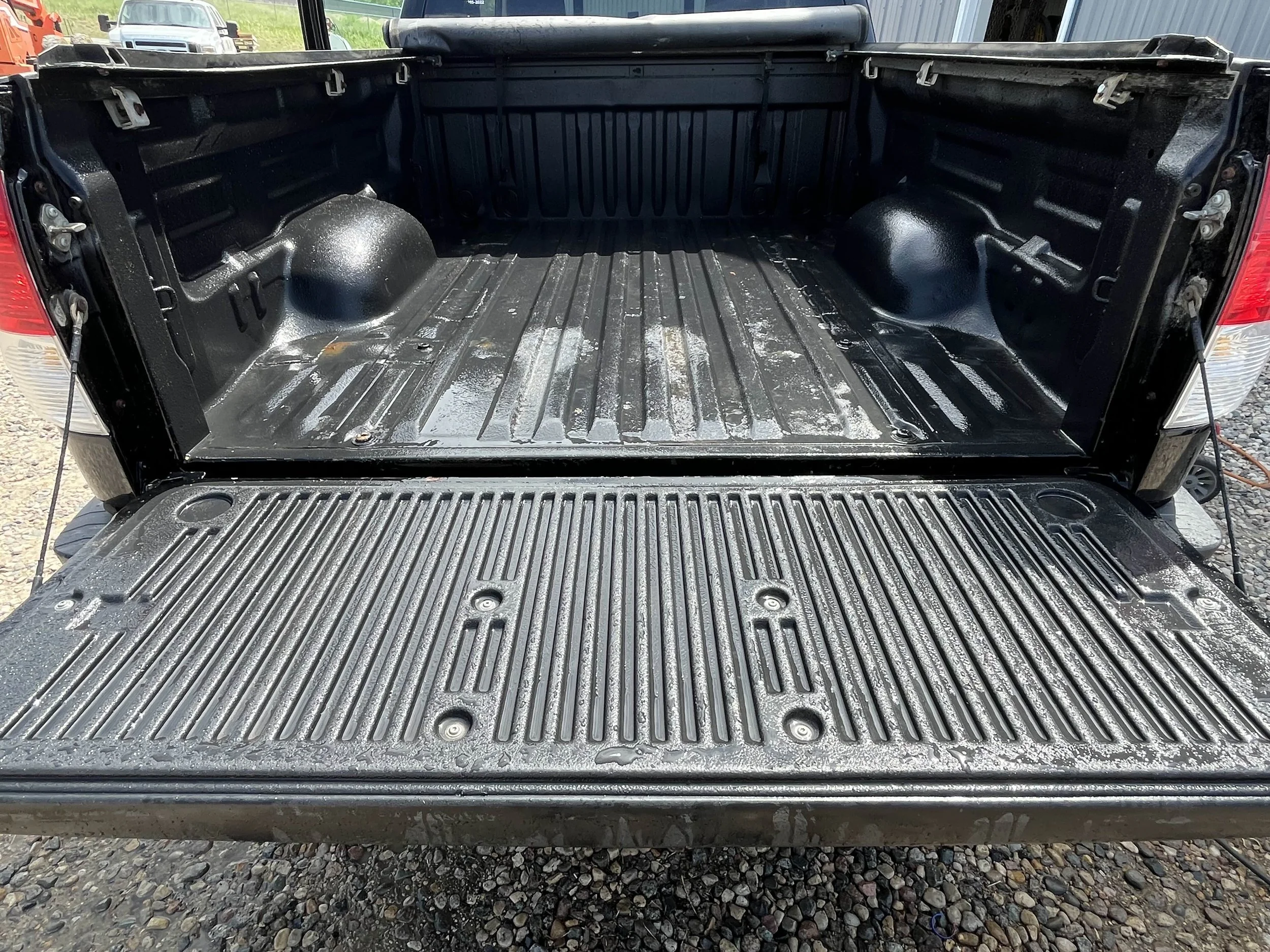 Empty pickup truck bed with a fold-down tailgate, showing a black painted interior and a black bed liner with grooves and textured surface.