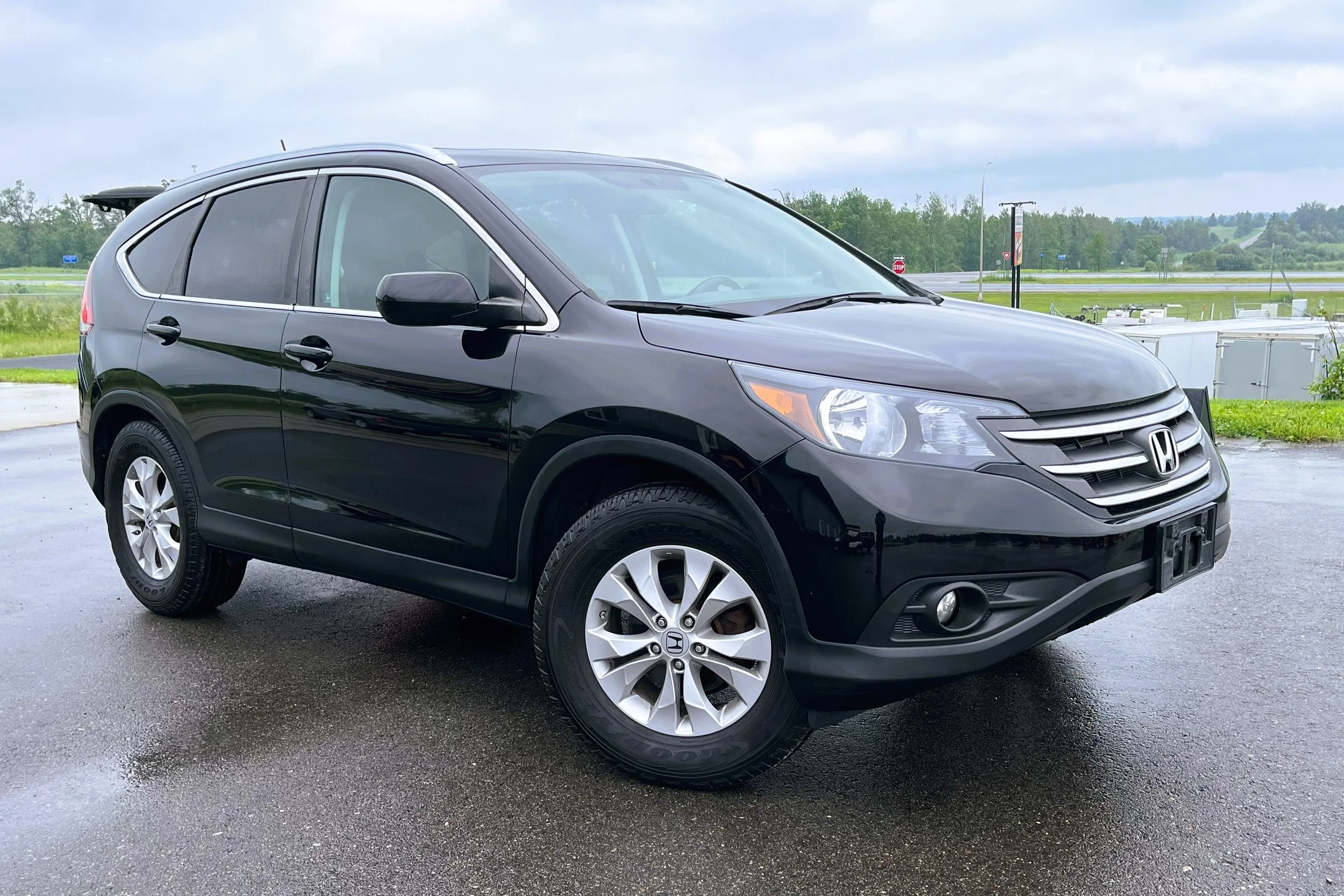 Black Honda CR-V parked outdoors on wet pavement, with green grassy fields and cloudy sky in the background.