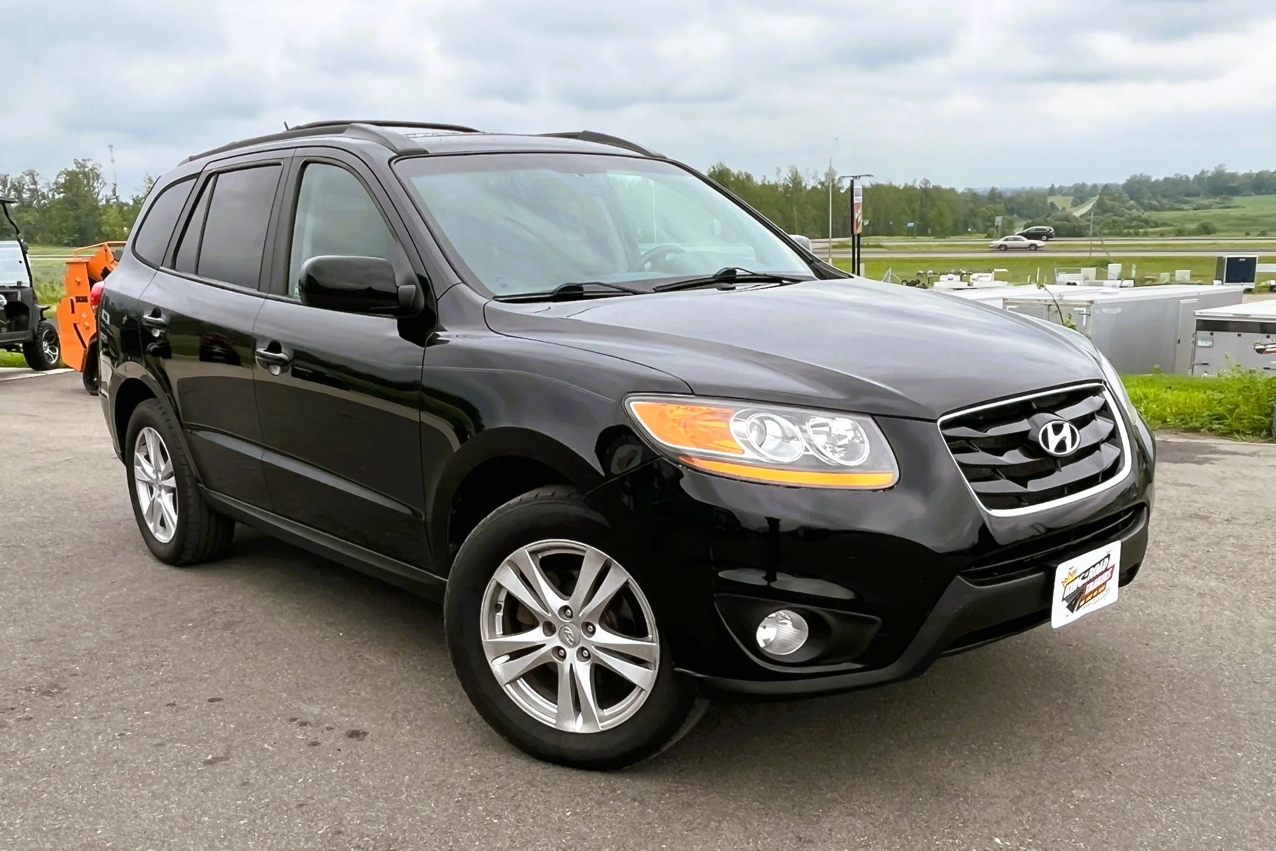 Black Hyundai SUV parked on a paved area outdoors with green fields and cloudy sky in the background.