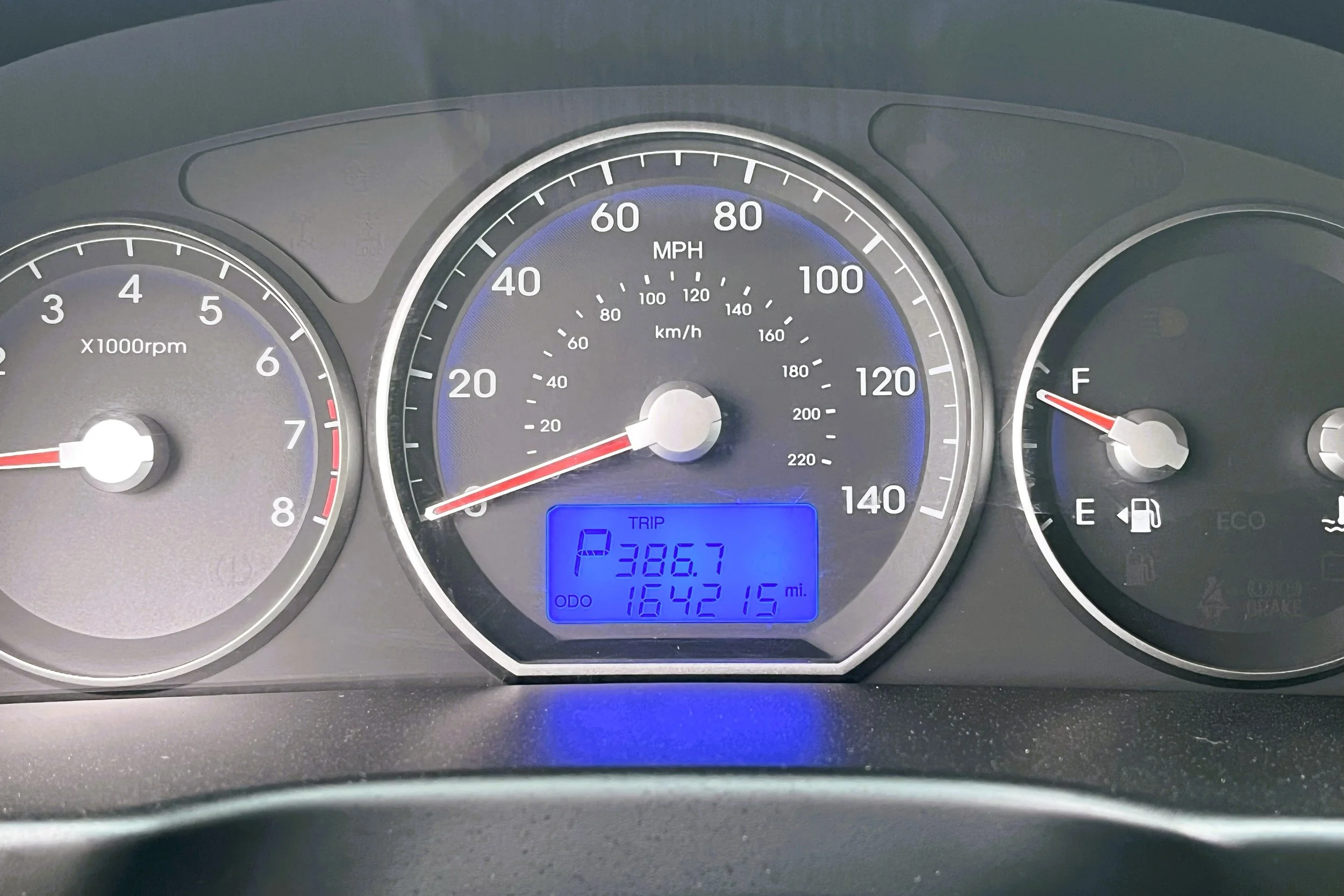 Car dashboard showing the speed at 0 MPH, the tachometer at about 8,000 RPM, fuel gauge near empty, and an odometer reading 1,642,15 miles. The display shows the vehicle in park with a trip distance of 386.7 miles.