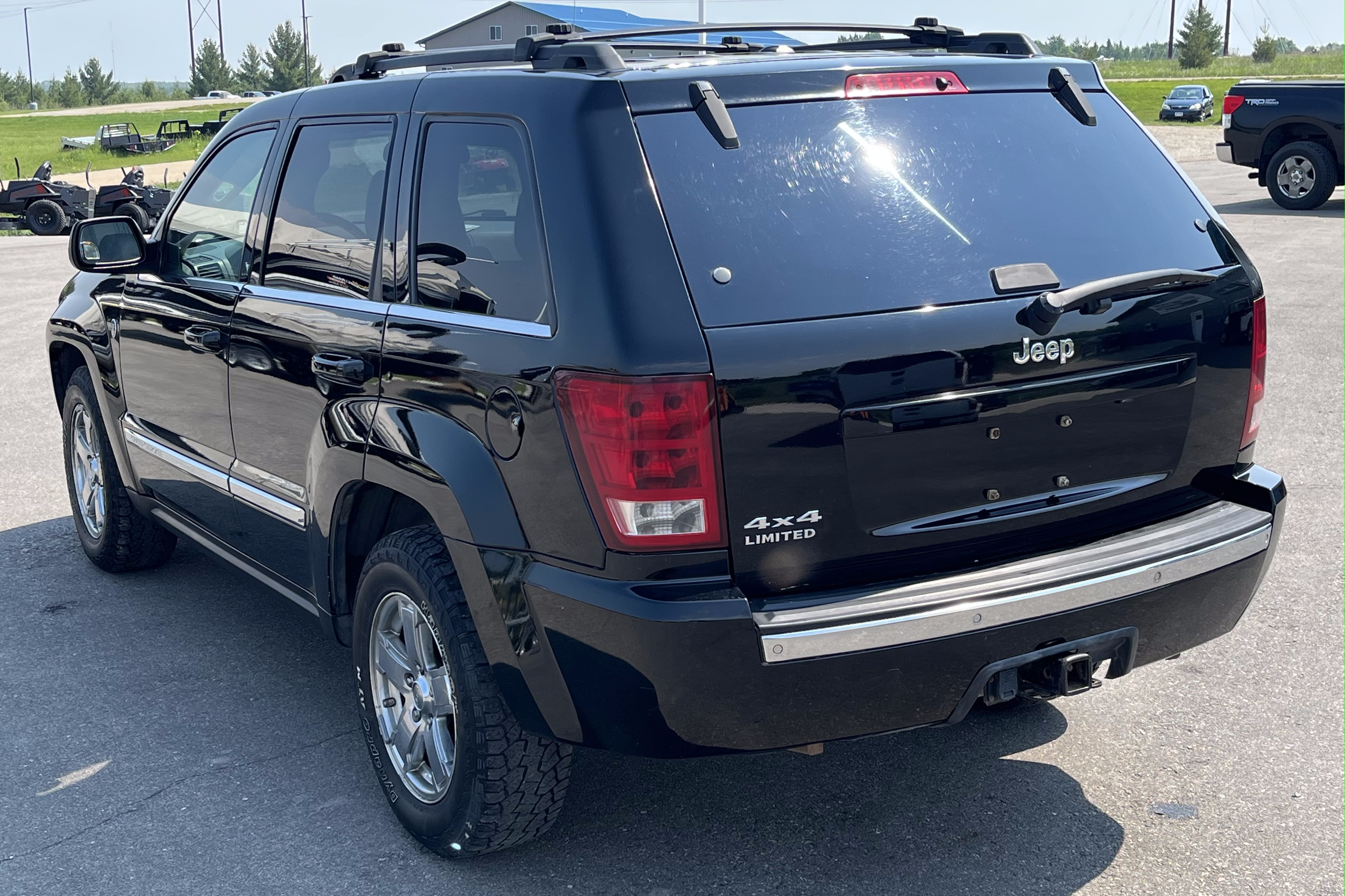 Black Jeep SUV parked outdoors on a paved lot, with a trailer hitch and 4x4 Limited badge on the rear, and other vehicles in the background.