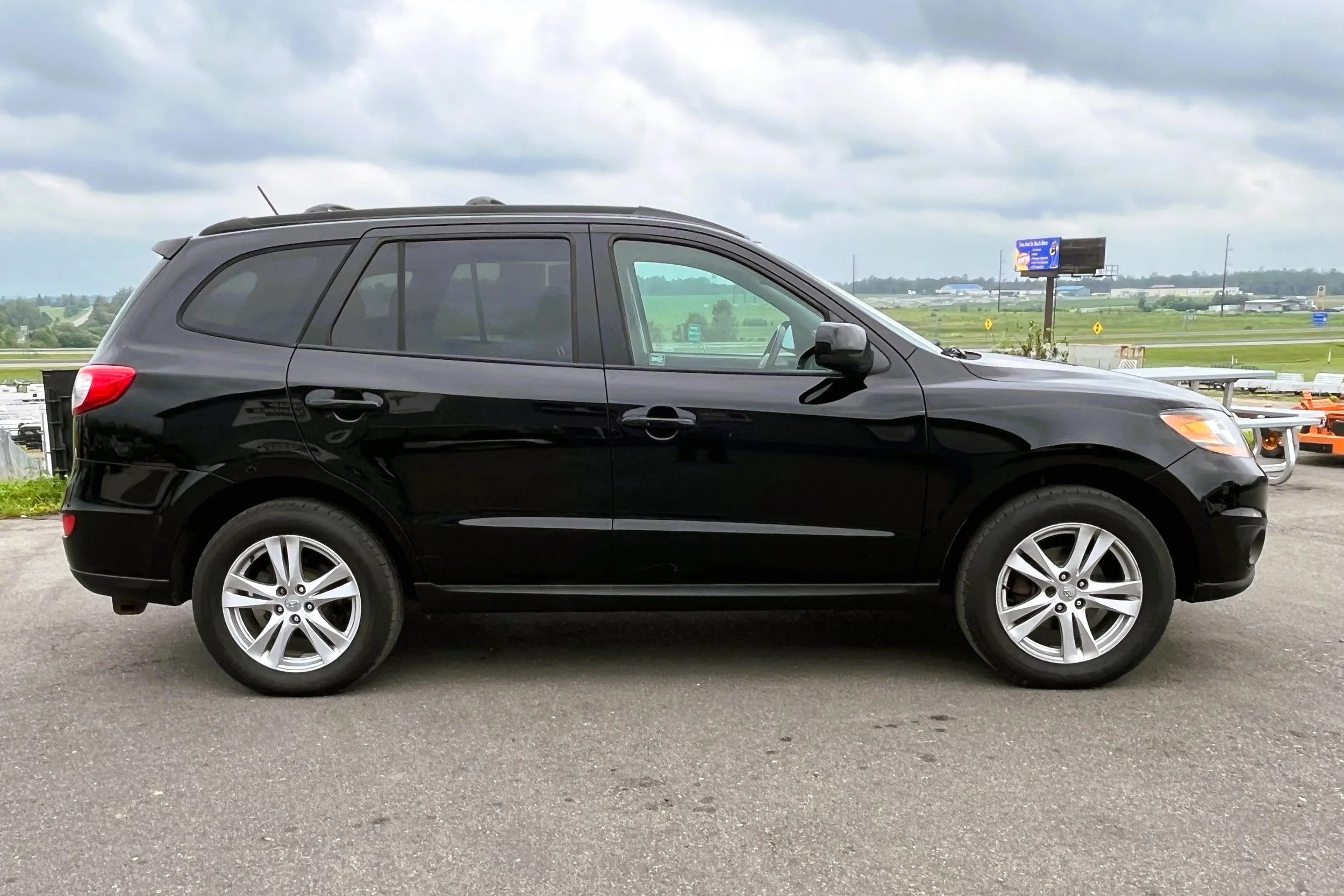 Side view of a black compact SUV parked on asphalt with green fields, a highway, and billboards in the background under cloudy skies.
