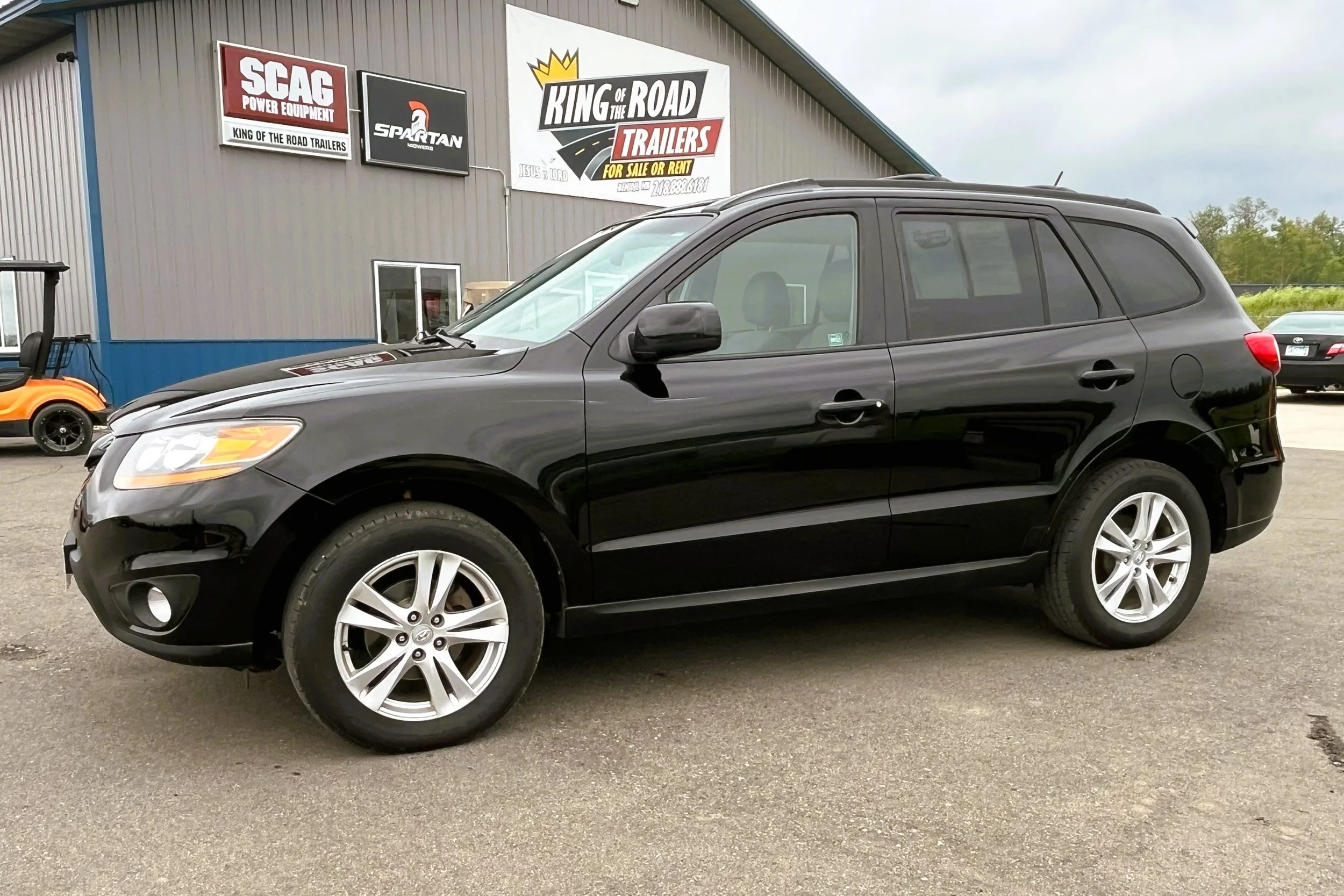 Black mid-sized SUV parked in front of a building with car dealership signs, including 'King of the Road Trailers' and 'SPARTAN.'