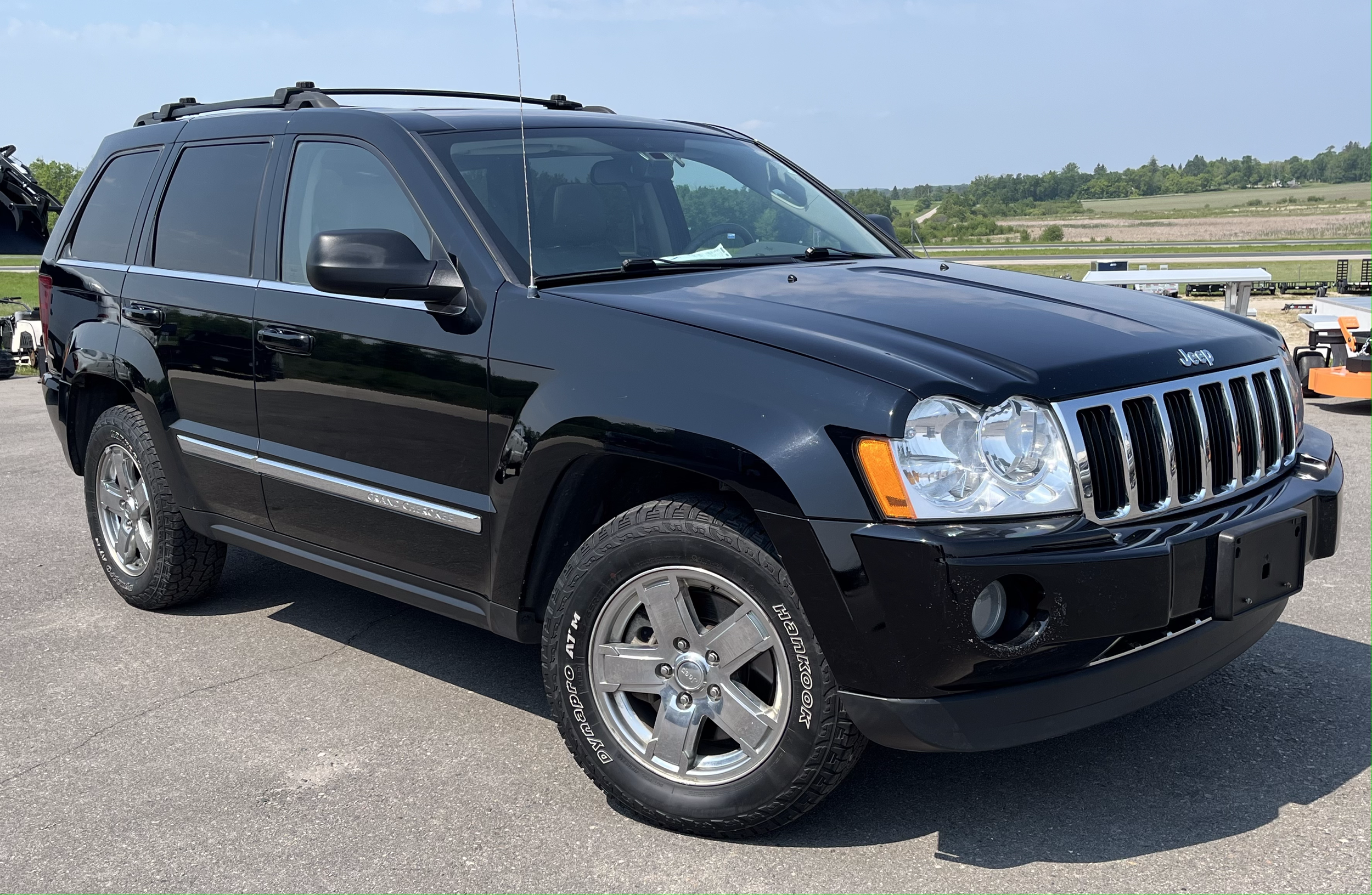 Black Jeep SUV parked outdoors on pavement with a grassy hill and blue sky in the background.