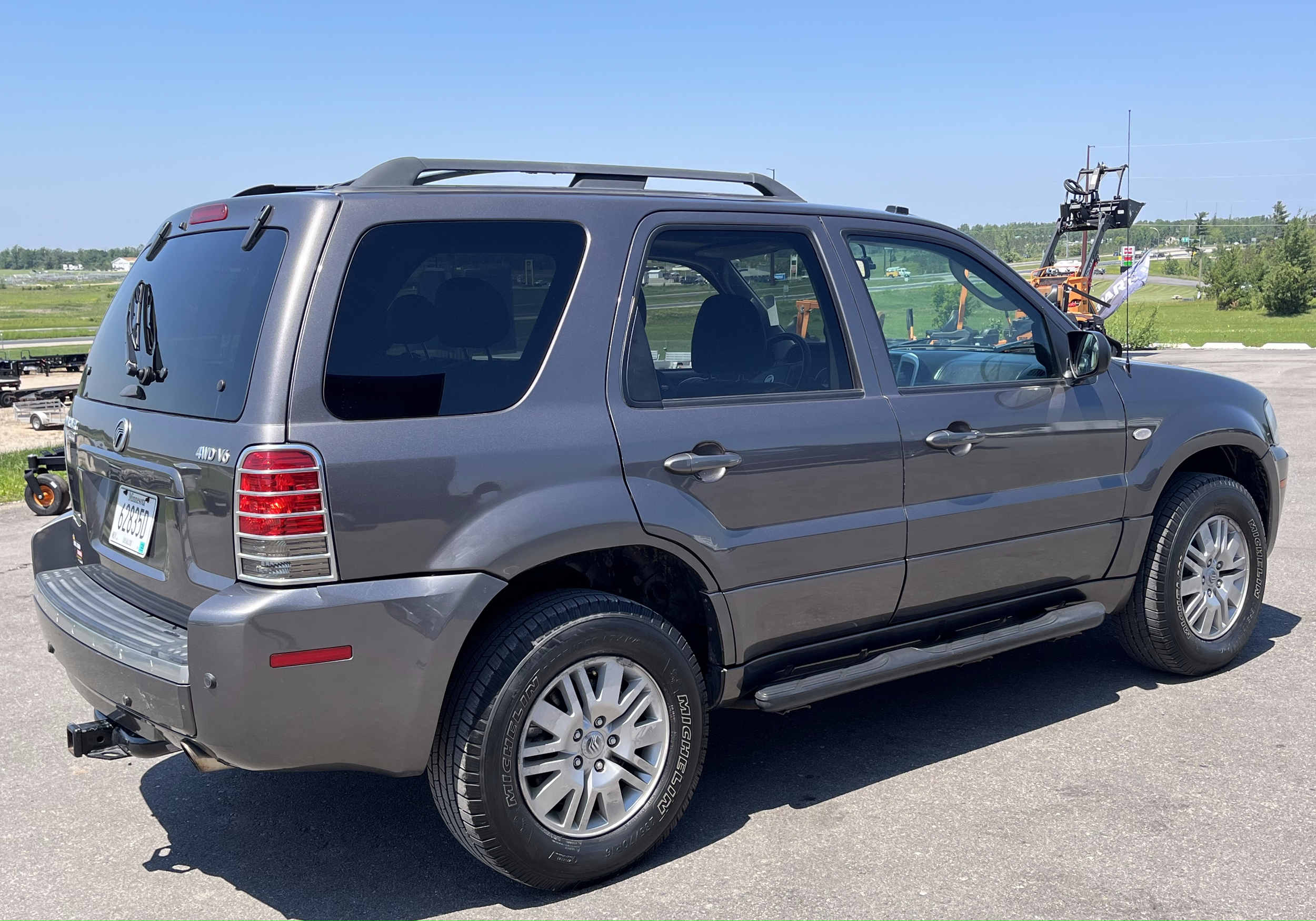 Gray SUV with four doors parked outdoors on paved surface, with green fields and machinery in the background.