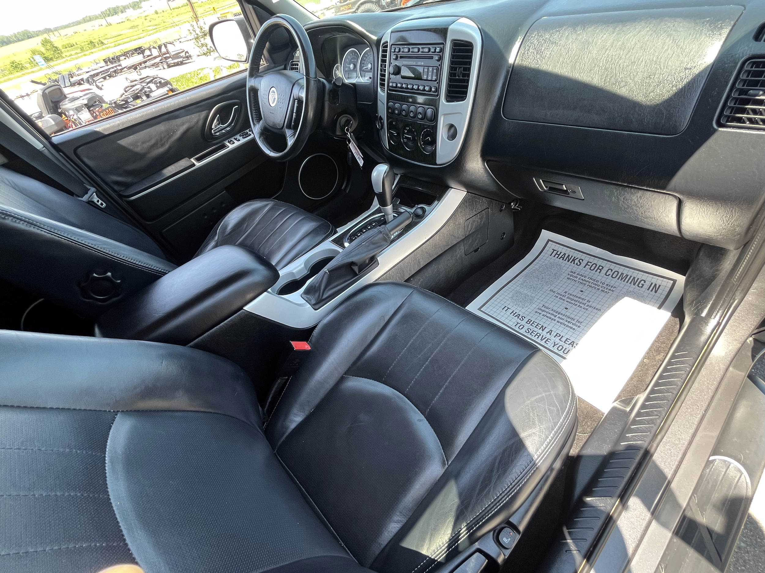 Interior of a vehicle showing the front seats, dashboard, steering wheel, gear shift, and a paper on the passenger side floor mat that reads "Thanks for coming in."