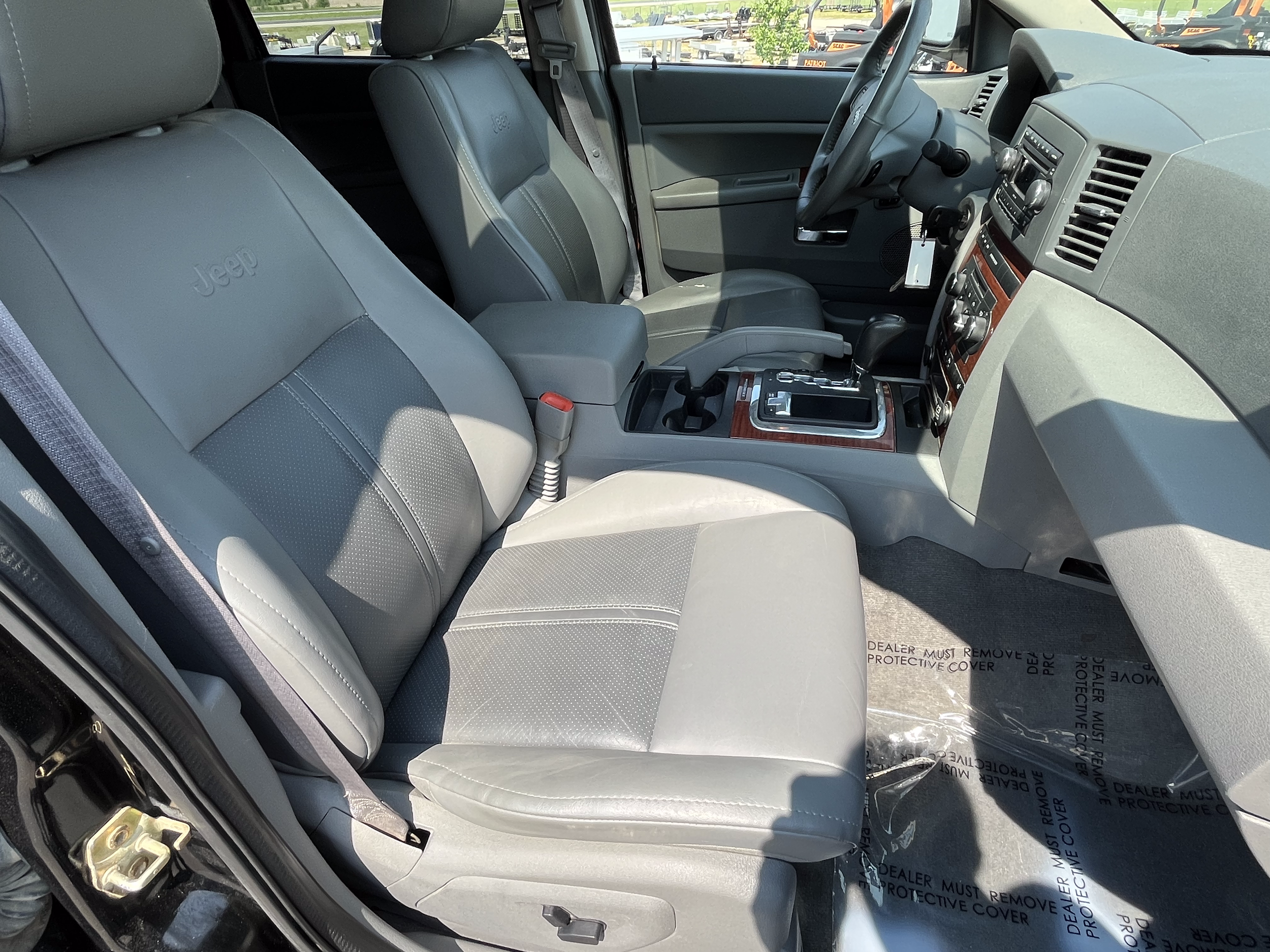 Interior of a vehicle showing the front two seats with gray leather upholstery, center console, dashboard, and gear shift. The front passenger seat is visible, with sunlight casting shadows inside.
