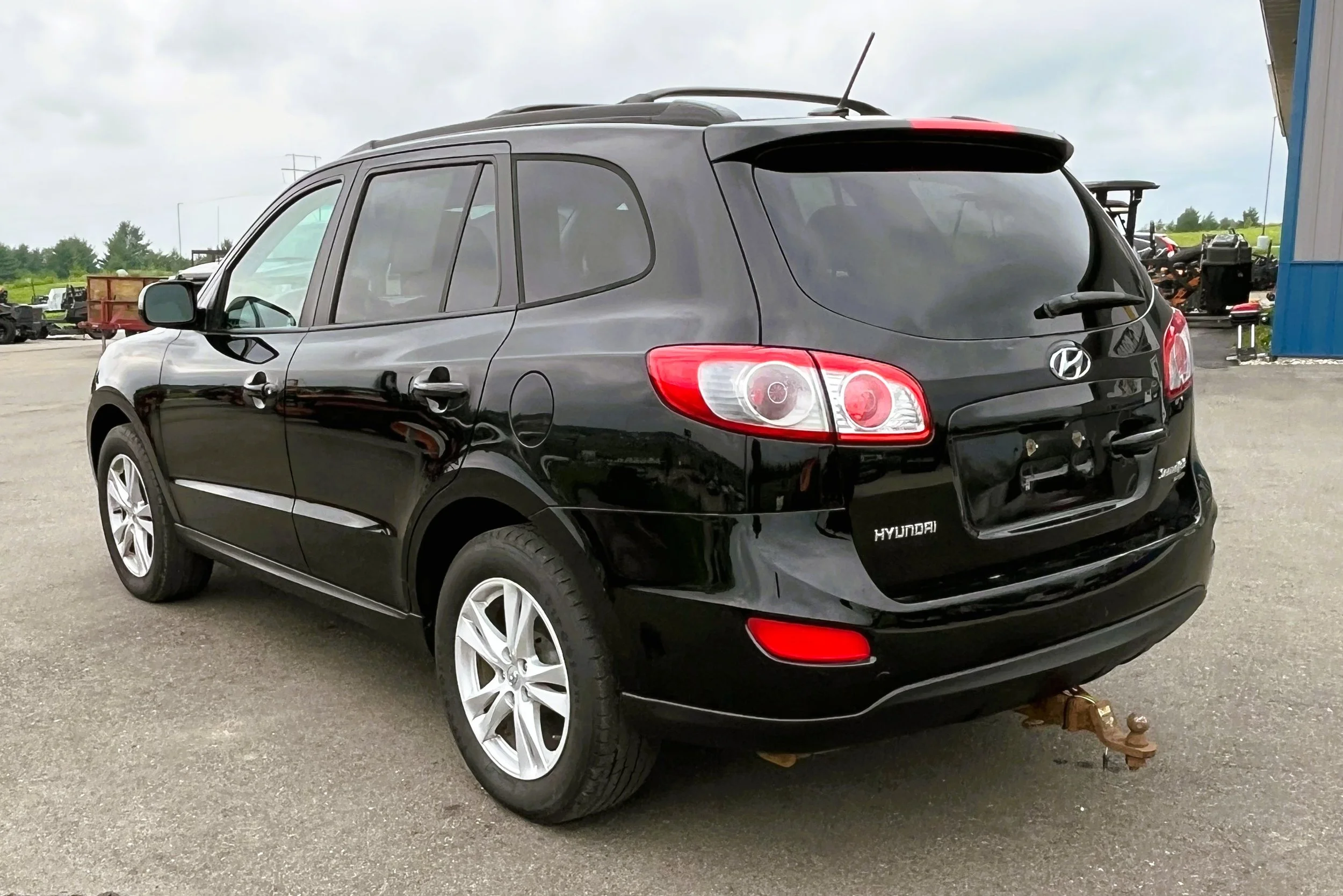 Black Hyundai SUV parked outdoors on a gray paved surface, with a blue building and machinery in the background.