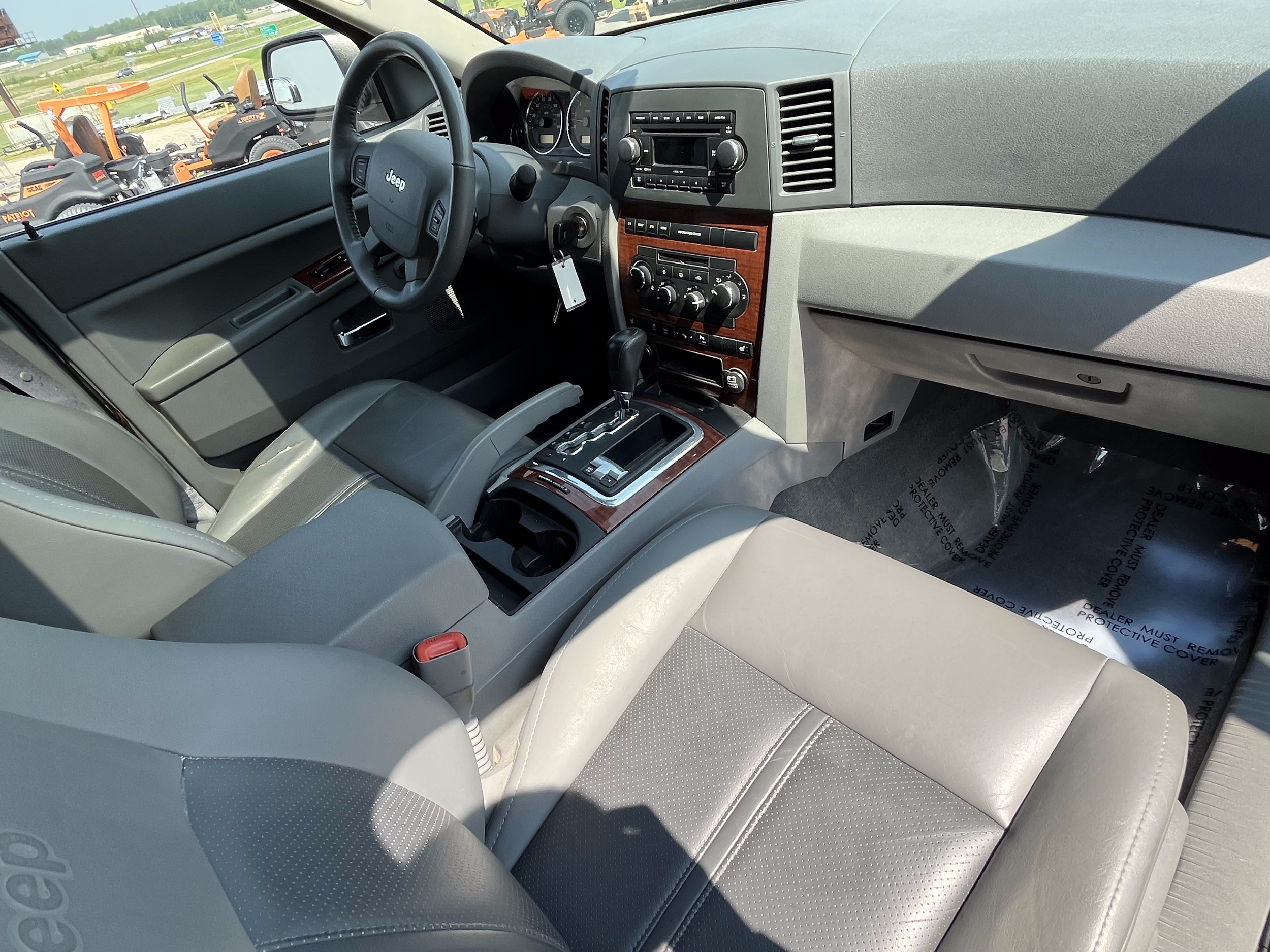 Interior of a gray Jeep vehicle showing dashboard with wood paneling, gear shift, steering wheel, and front seats, with outdoor vehicles visible through the windshield.