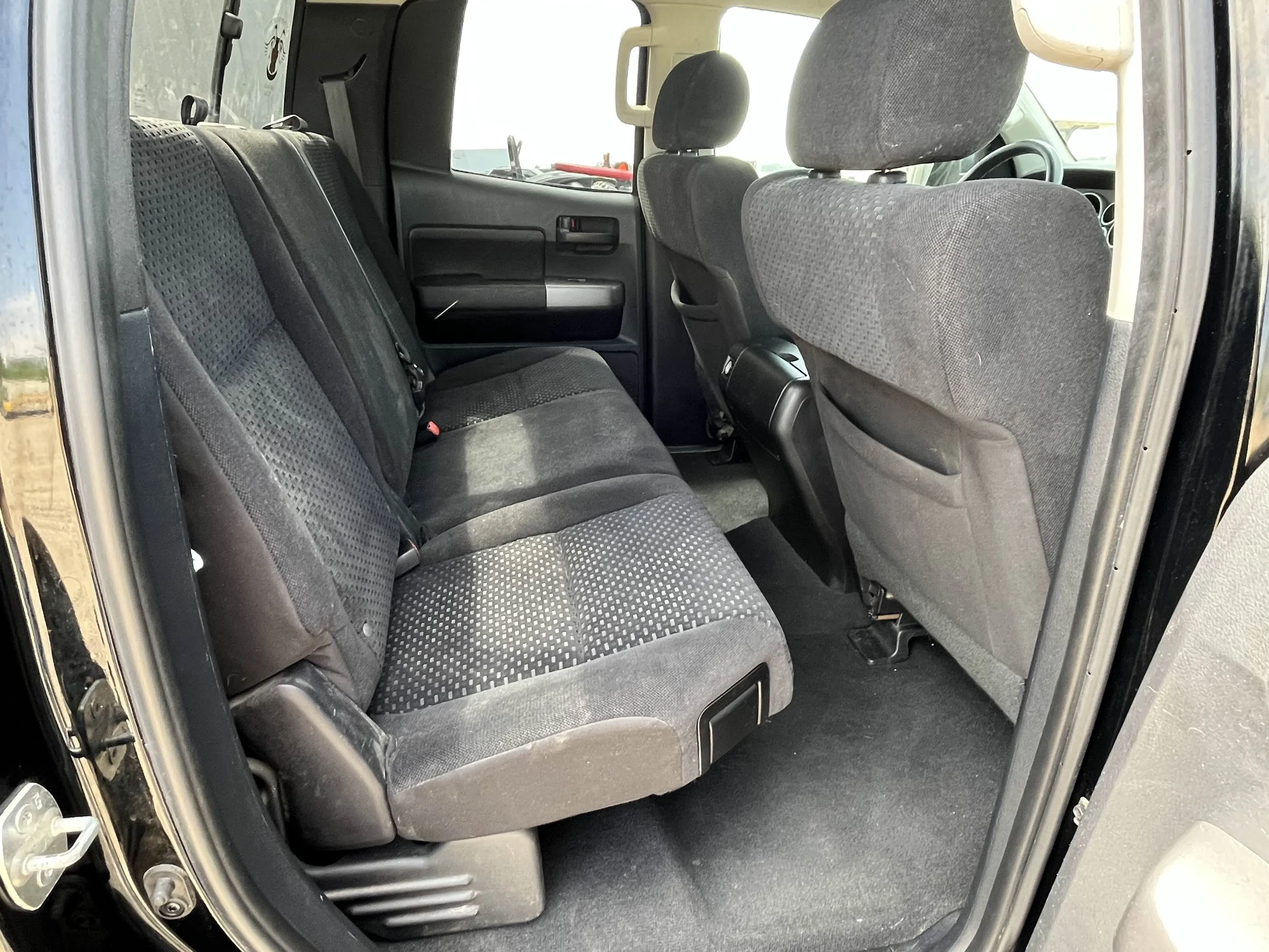 Rear interior of a vehicle showing the back seats, which are covered in dark fabric, and the front seats, which are also covered in dark fabric with headrests. The floor is black carpet.