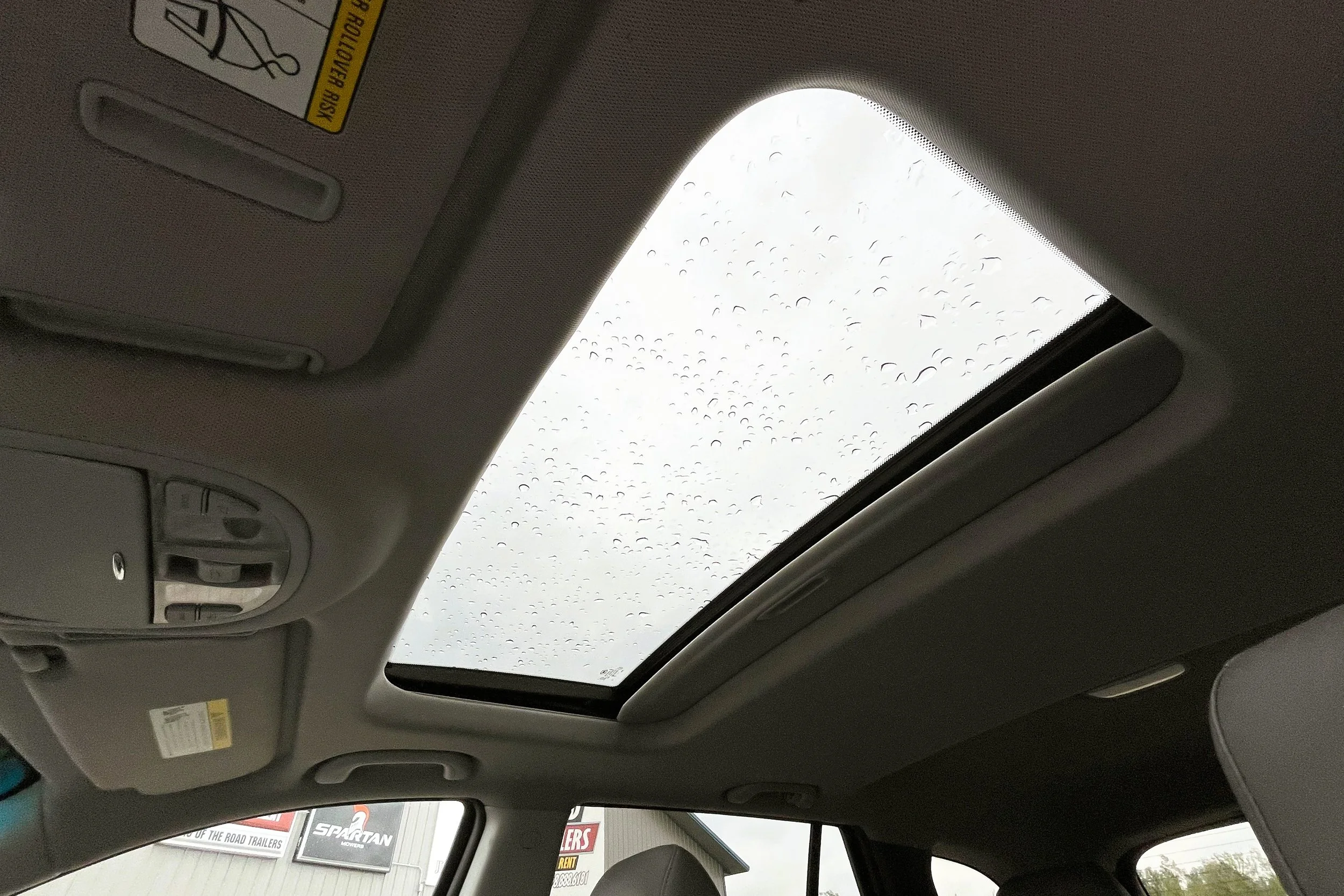 Car interior showing sunroof with raindrops on glass, side window, and ceiling controls.