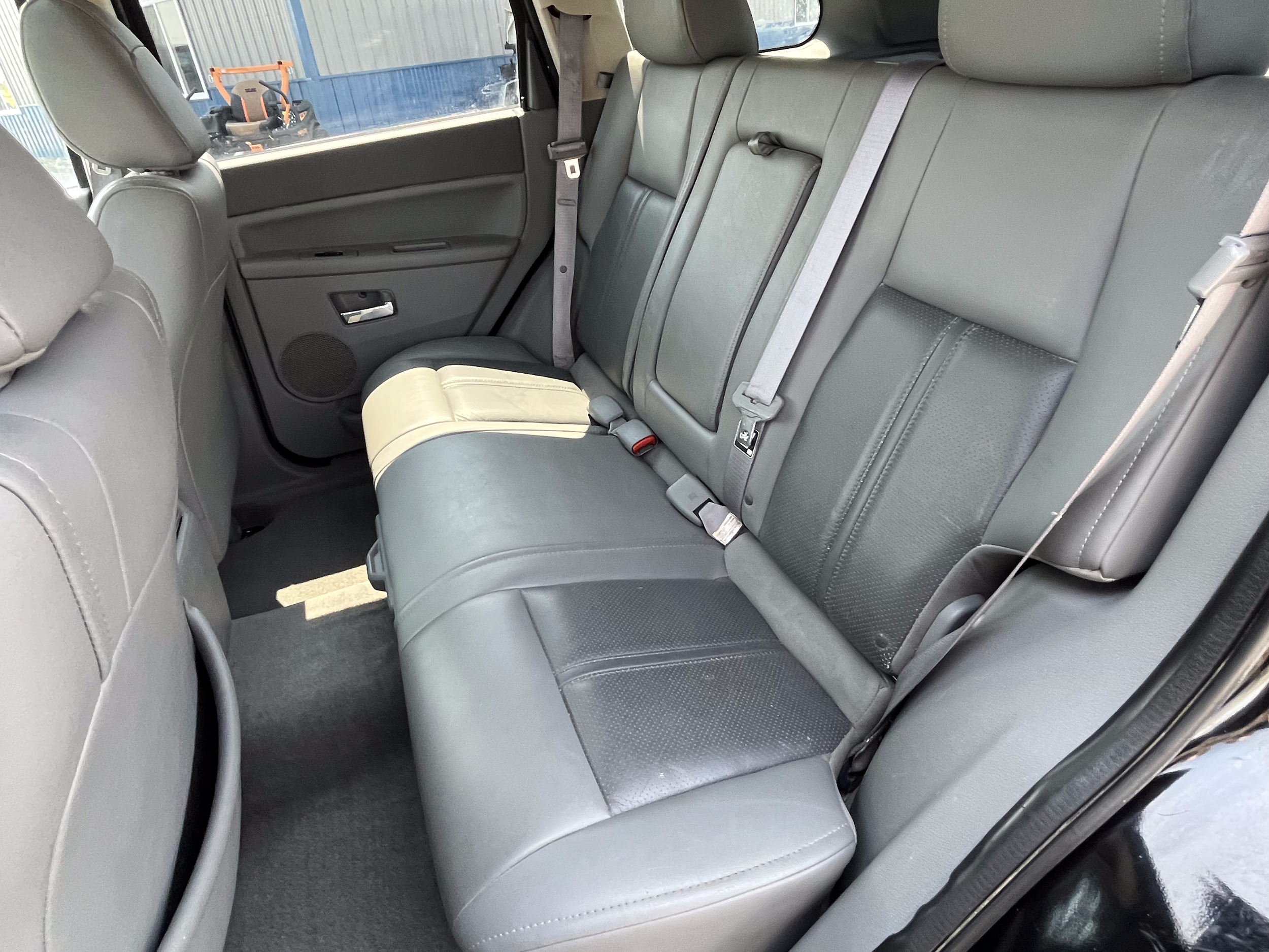The interior of a vehicle showing a rear seat with three seat belts, gray upholstery, and a fold-down center armrest.
