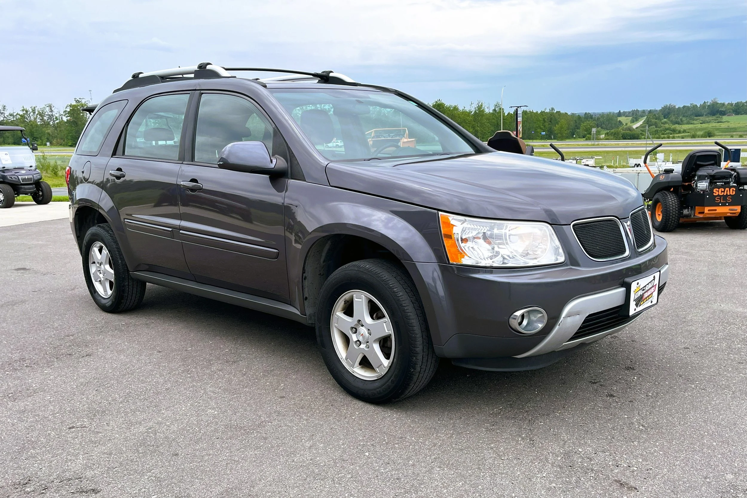 Gray Saturn Vue SUV parked on asphalt with a grassy field and trees in the background, alongside other vehicles and equipment.