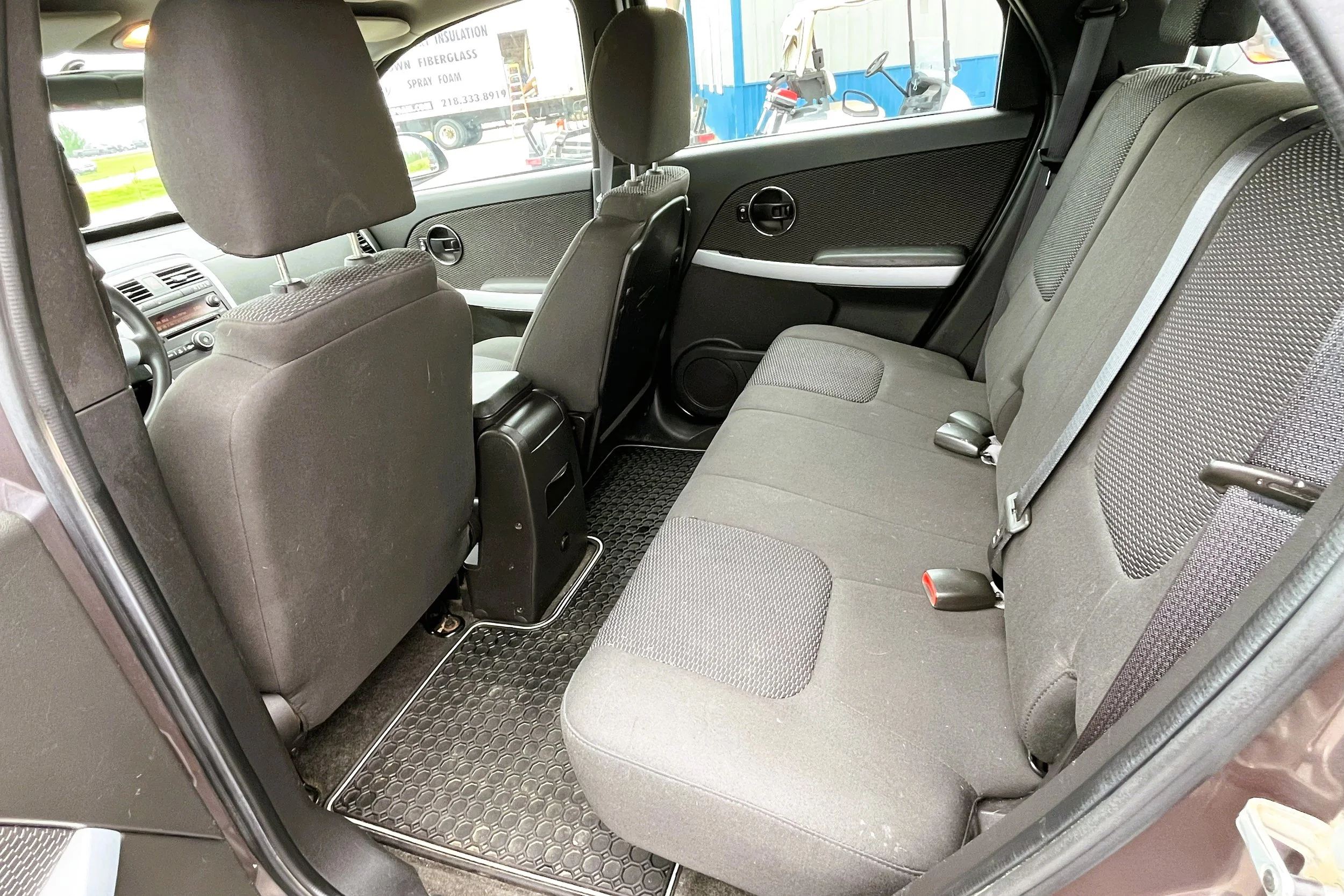 Interior of a vehicle showing gray fabric back seats, headrests, seat belts, and black rubber floor mats.