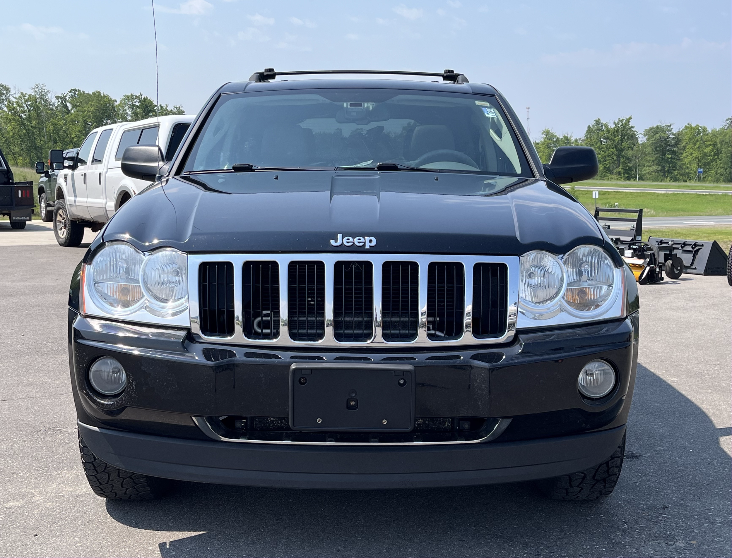 Front view of a black Jeep SUV parked outdoors with other vehicles and equipment in the background on a sunny day.