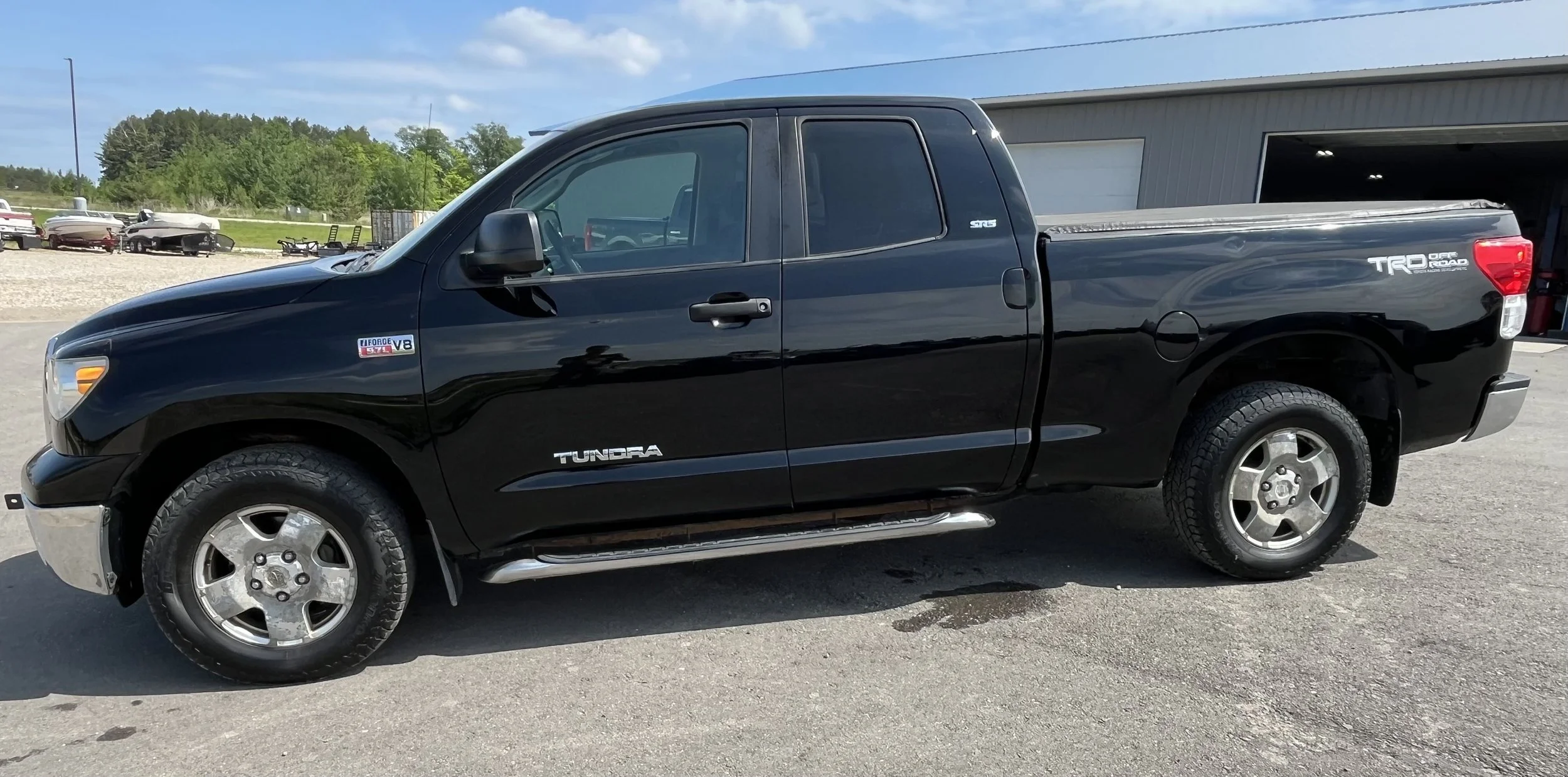 Black Toyota Tundra pickup truck parked on a gravel lot with boats in the background and a warehouse building nearby.