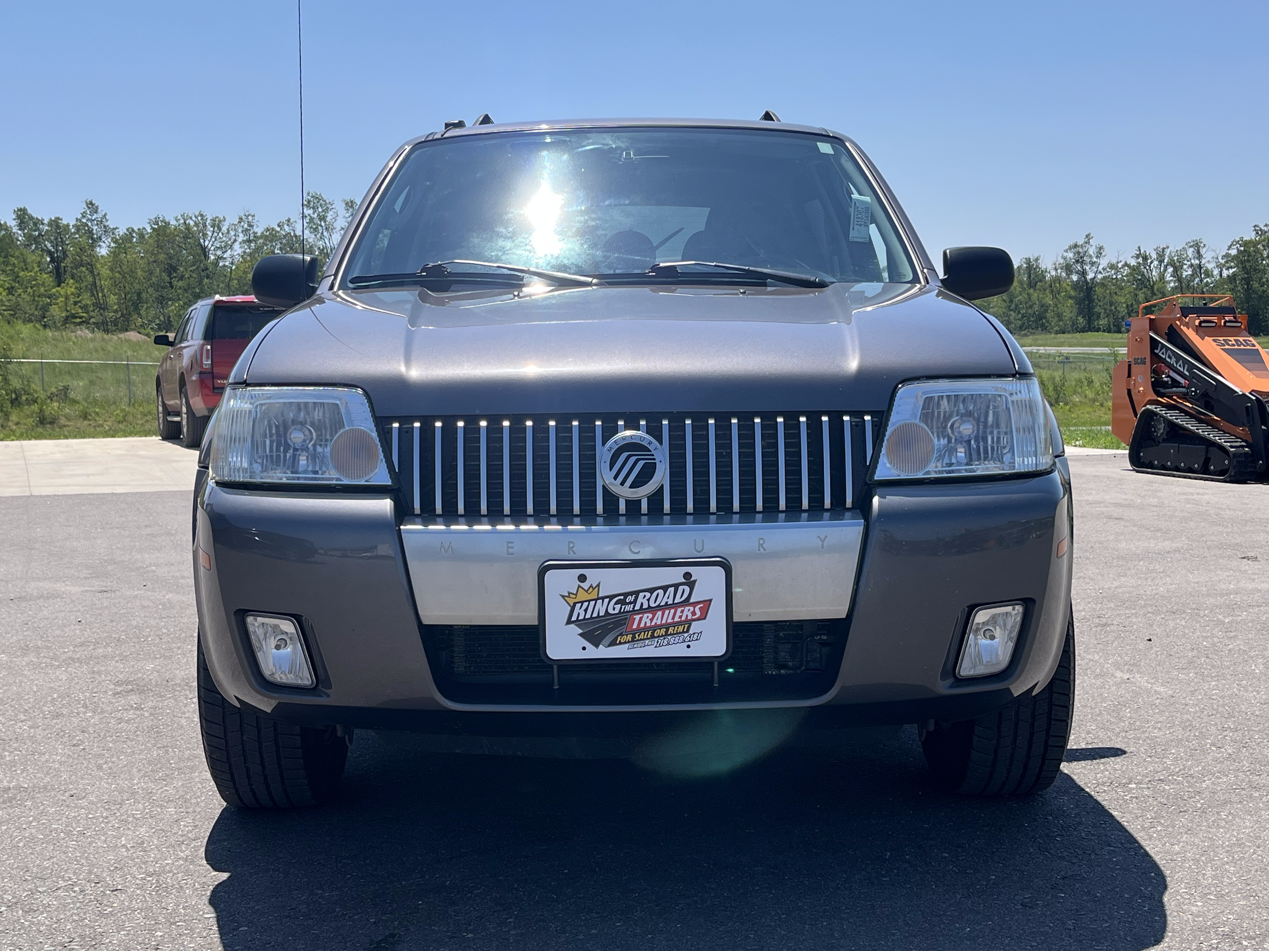 Front view of a gray Mercury SUV parked on pavement with a dealership license plate and a background of trees, another vehicle, and construction equipment.
