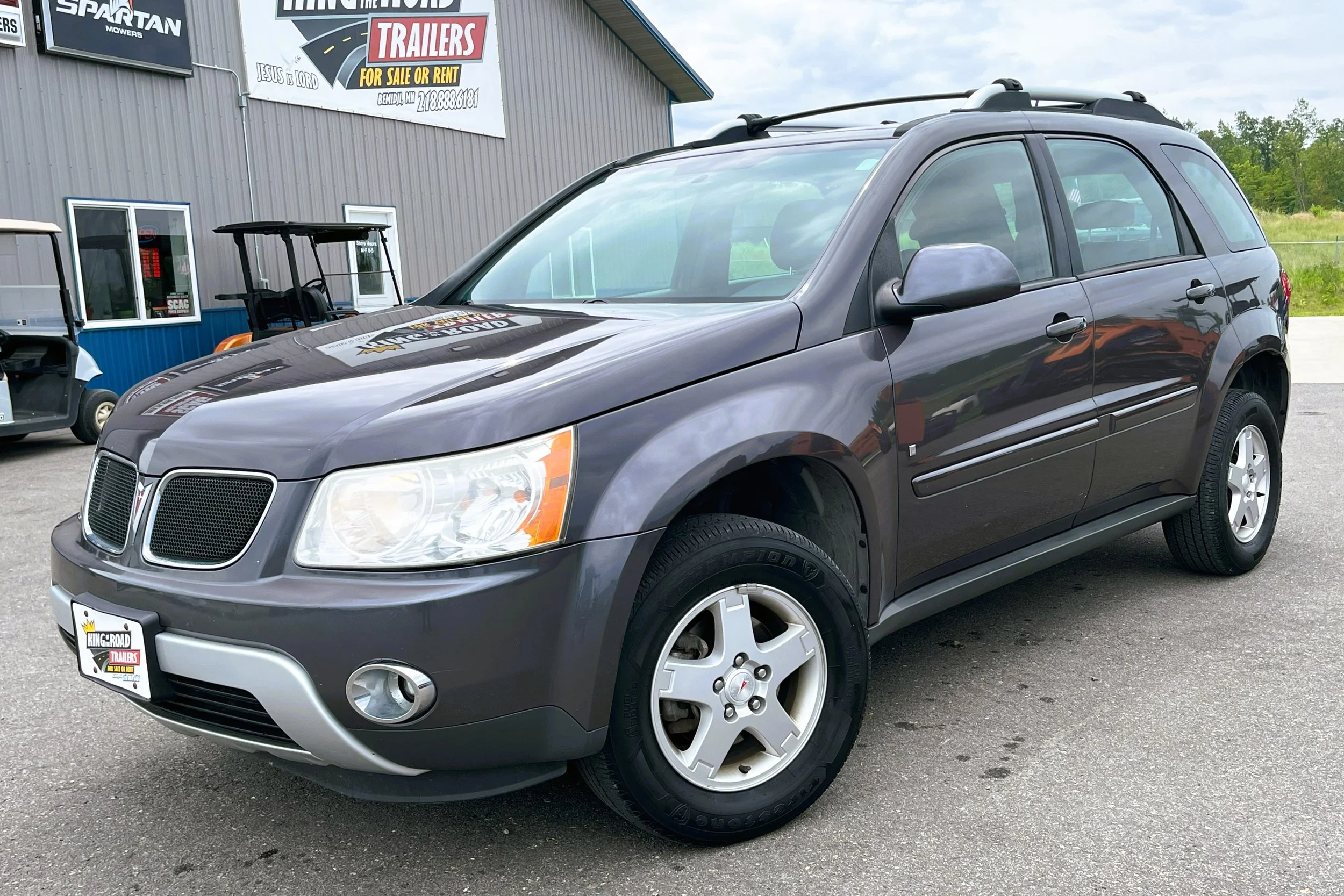 A dark gray Pontiac SUV parked at a dealership lot with a sign for trailers for sale or rent in the background.
