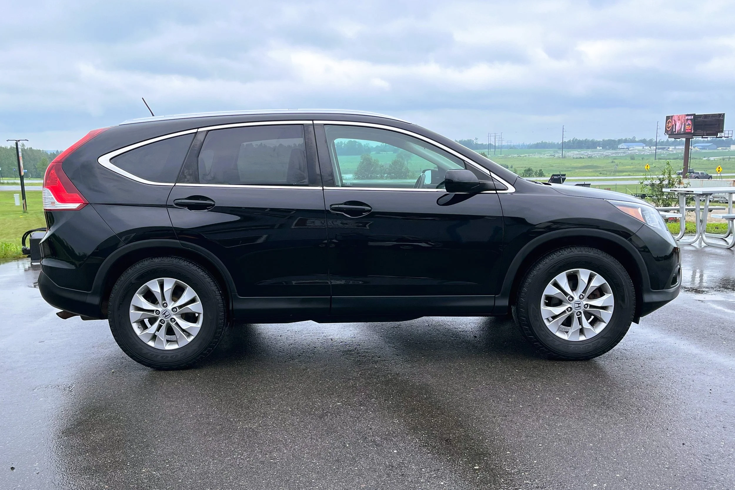 Side view of a black Honda CR-V parked outdoors on wet pavement with a grassy landscape and cloudy sky in the background.