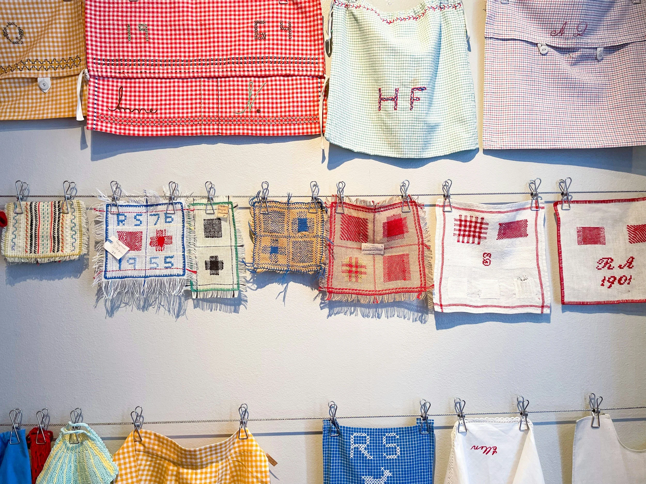 Display of embroidered, checkered cloths and small fabric pieces with various designs hanging on a wire wall display.