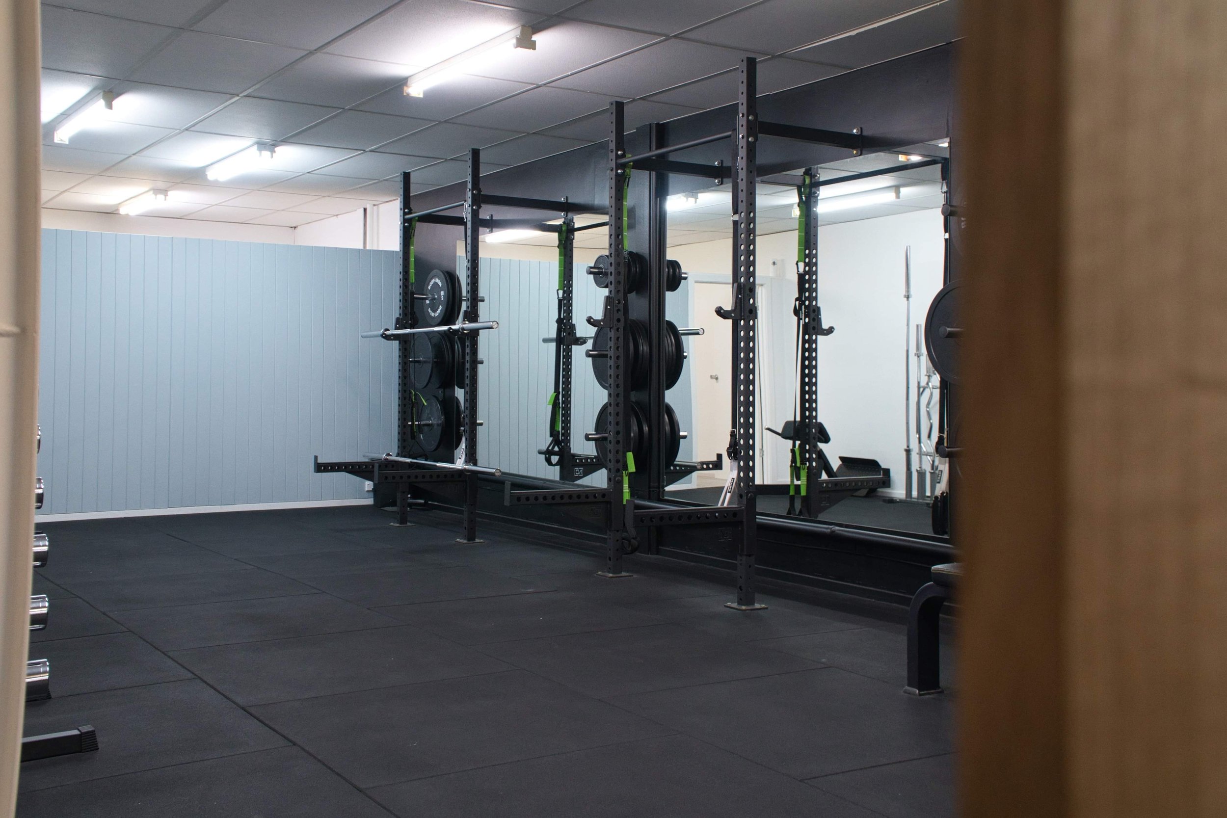 Empty gym with black weightlifting racks, barbells, and weight plates, mirrored wall, and rubber flooring.
