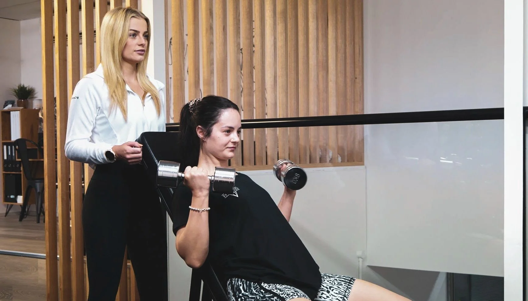 Woman in black shirt and zebra-print shorts doing seated bicep curls with dumbbells while a trainer stands behind her at a gym.