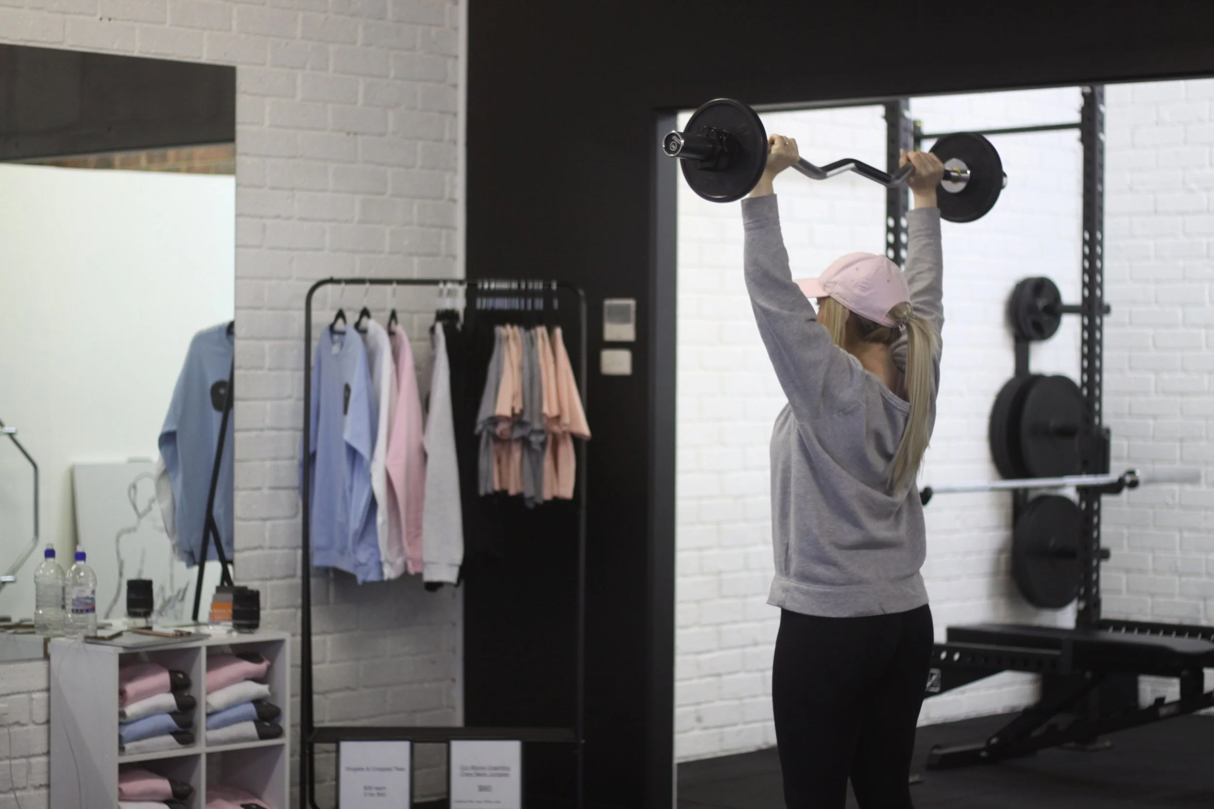 A woman in a pink cap and gray sweatshirt lifting a barbell in a gym with white brick walls and black fitness equipment.