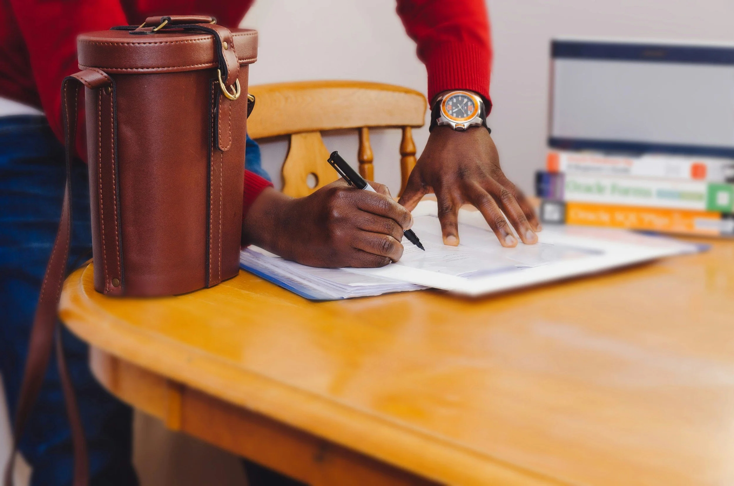 A person is writing in a notebook on a wooden table with documents and books, wearing a wristwatch and a red sweater. A brown leather bag is on the table.